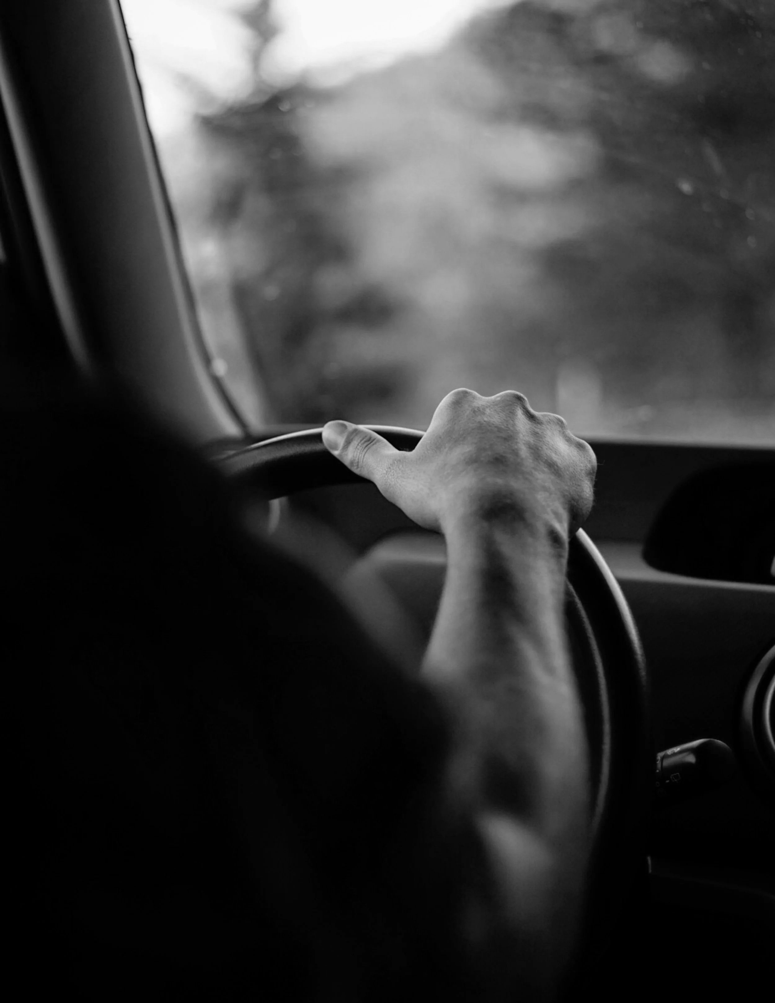 Black and white photo of a person driving a vehicle, focusing on their hand on the steering wheel, with the view through the windshield showing a blurred outside scene | Rental Studio for Film, Photography and Events | Hembrug Zaandam | Greater Amste