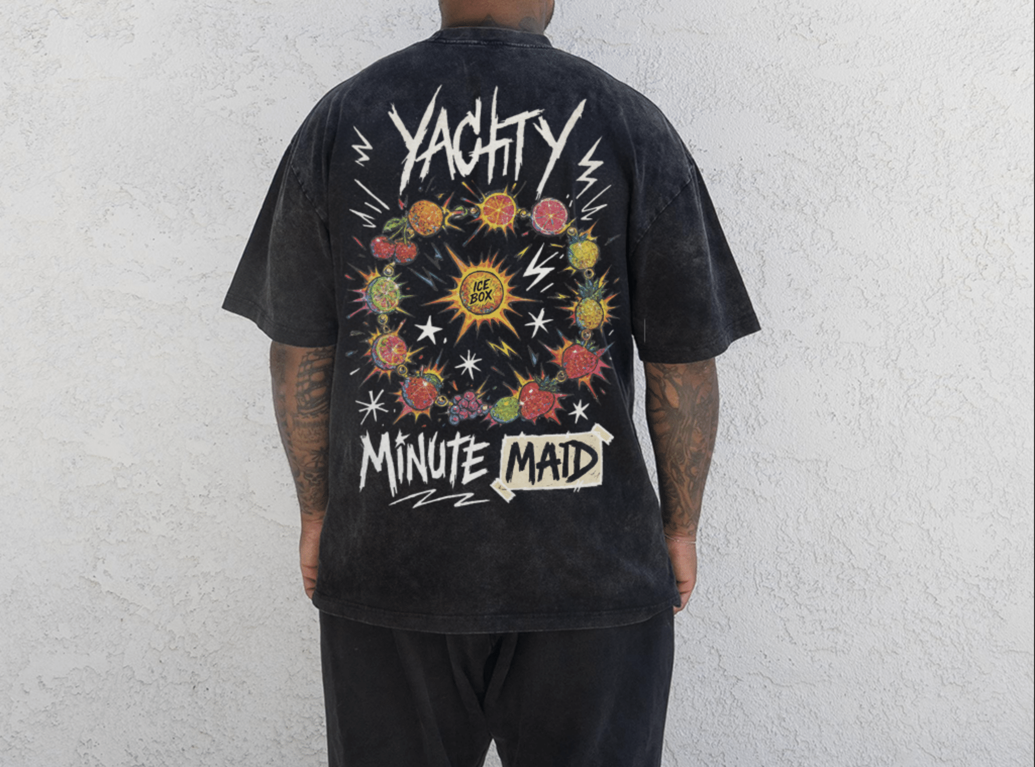 Back view of a person wearing a black graphic T-shirt with colorful fruit and lightning bolt designs, and the words 'Yachty' and 'Minute Maid' printed on it, standing against a white textured wall.