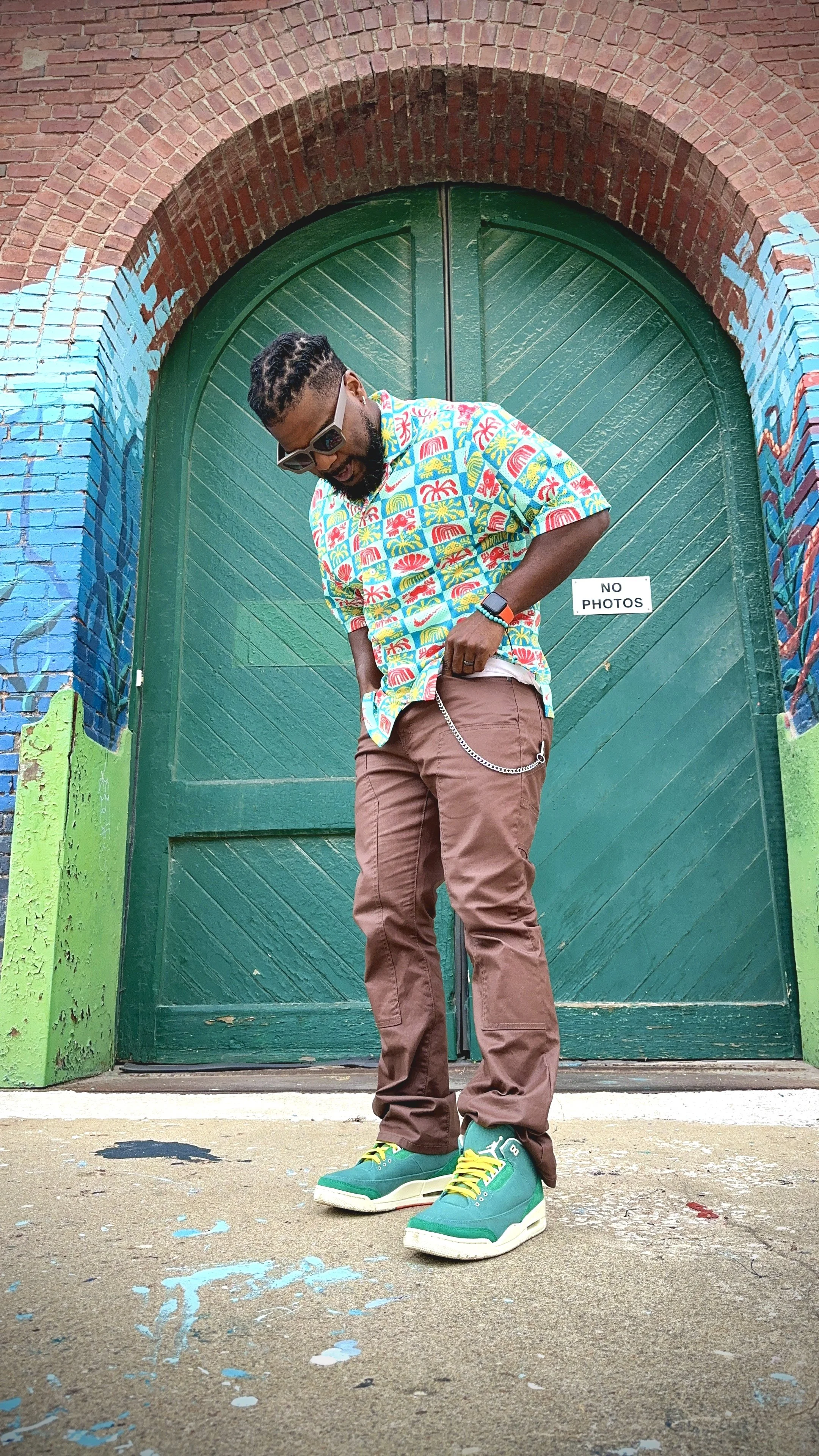 Man standing in front of a green door with a brick archway, wearing sunglasses, a colorful Hawaiian shirt, brown pants, and teal sneakers, looking downward, with a