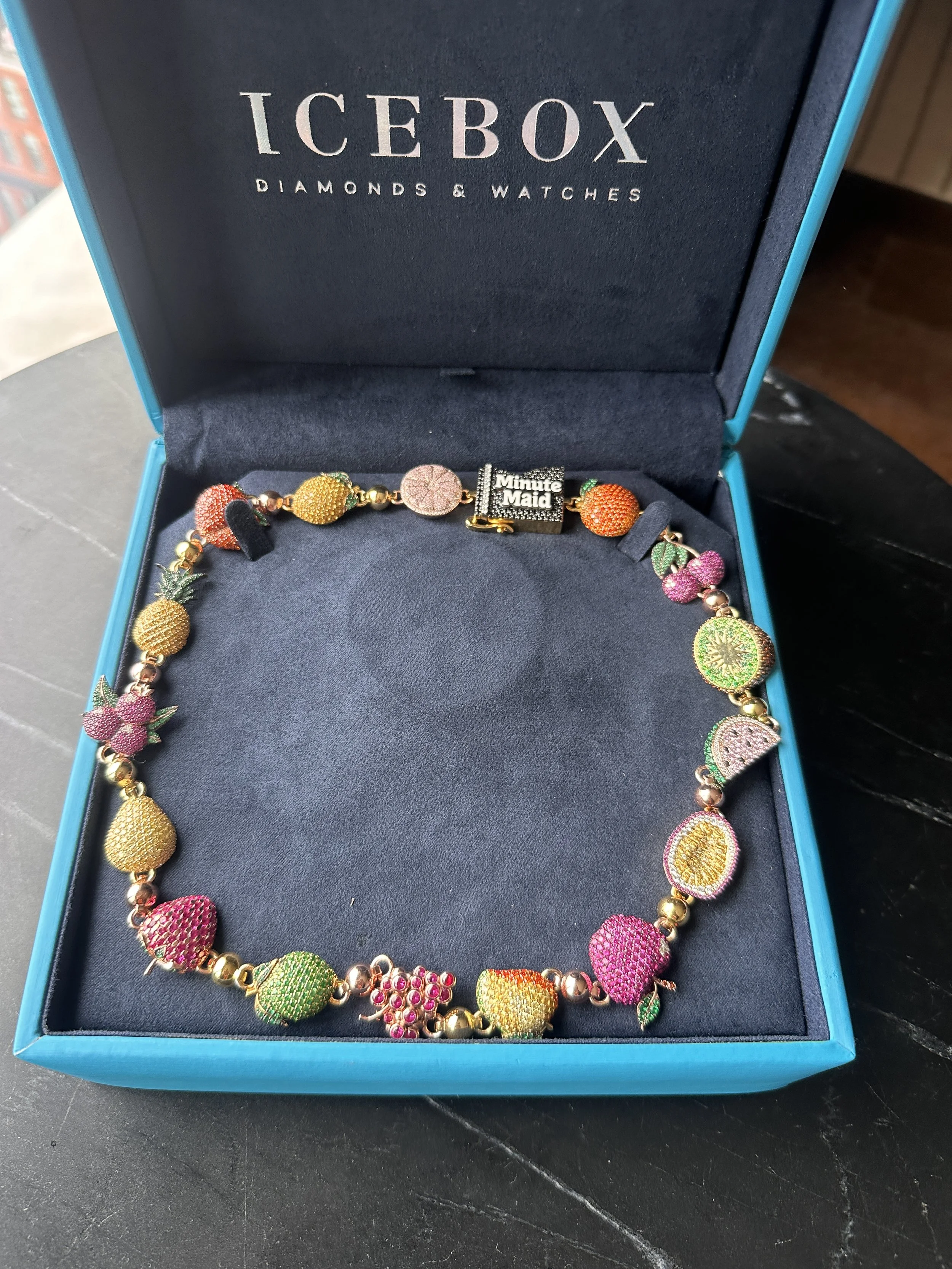 A colorful fruit-themed necklace with pineapple and berry-shaped pendants, displayed in an icebox jewelry box with a navy interior that says 'ICEBOX DIAMONDS & WATCHES' on the inside of the lid.