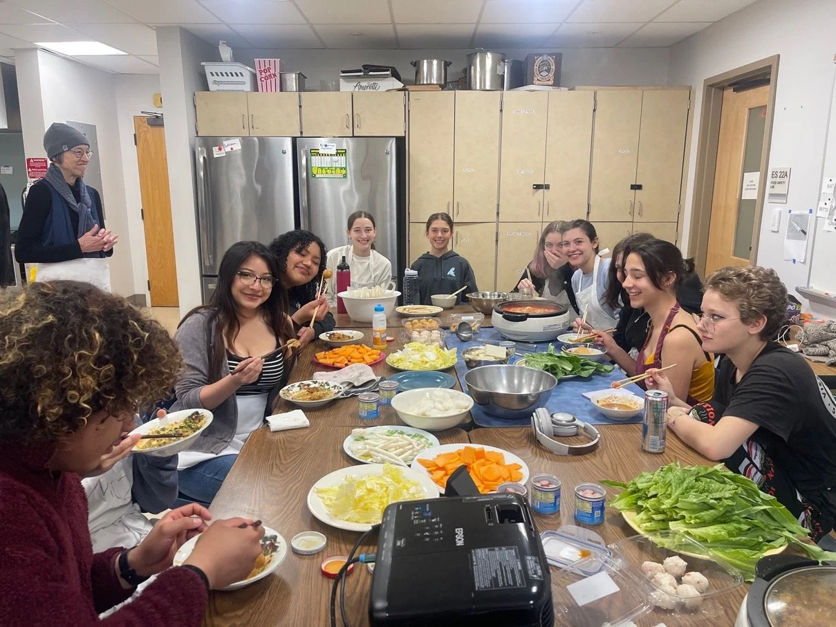 Group of young women gathered around a table preparing food in a kitchen, with various vegetables, bowls, and cooking utensils on the table.