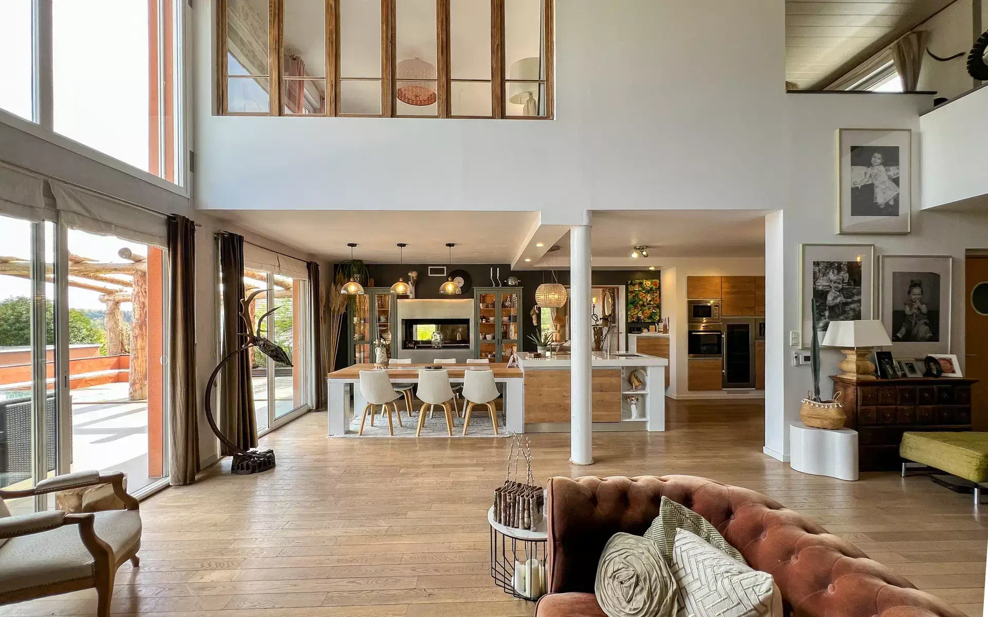 Modern open-concept living room and kitchen with wooden flooring, large windows, a brown sofa with decorative pillows, a dining table with chairs, and kitchen appliances.