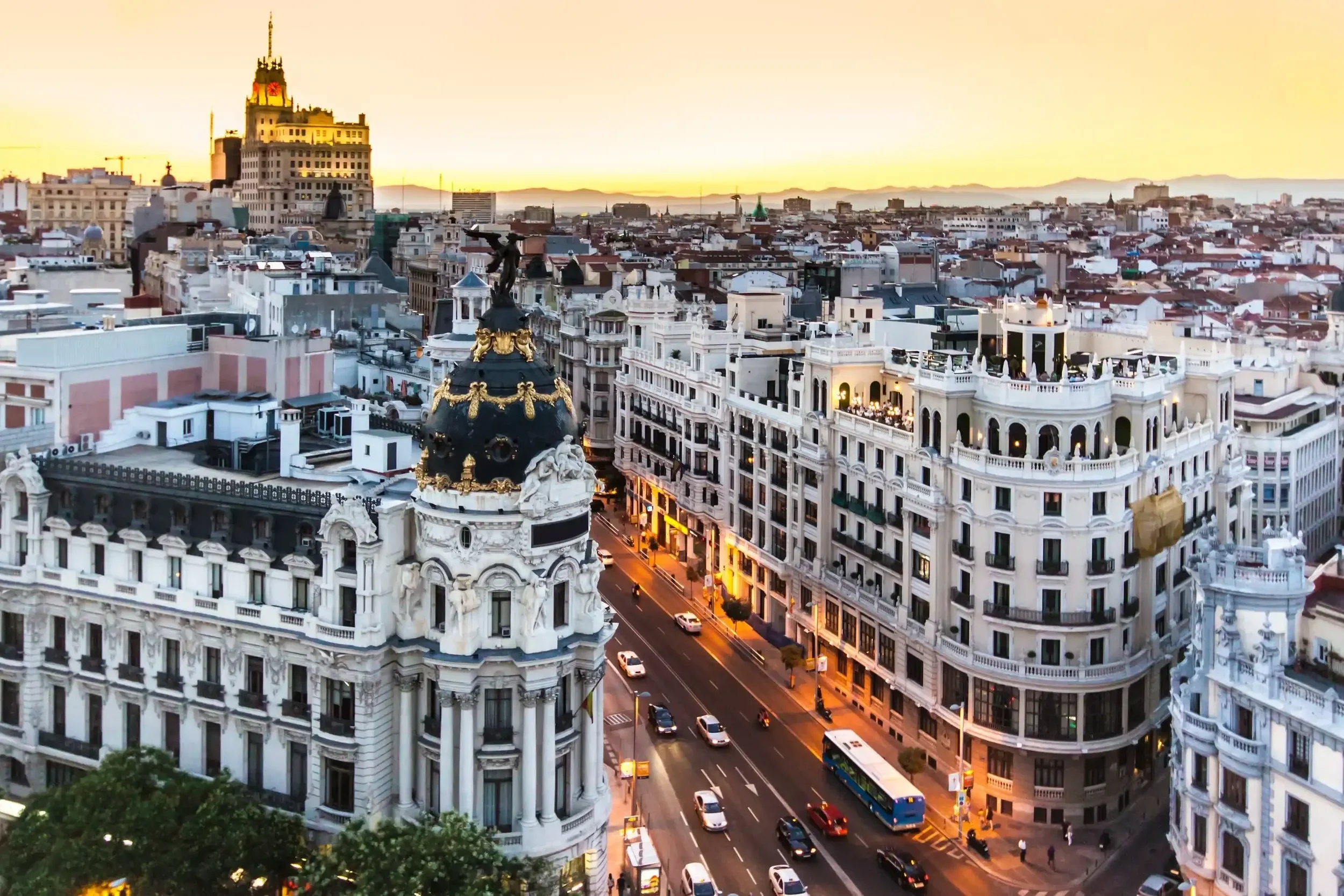 Cityscape at sunset featuring historic buildings with ornate architecture, busy streets, and the rooftop of a prominent building with a dome and golden decorations.