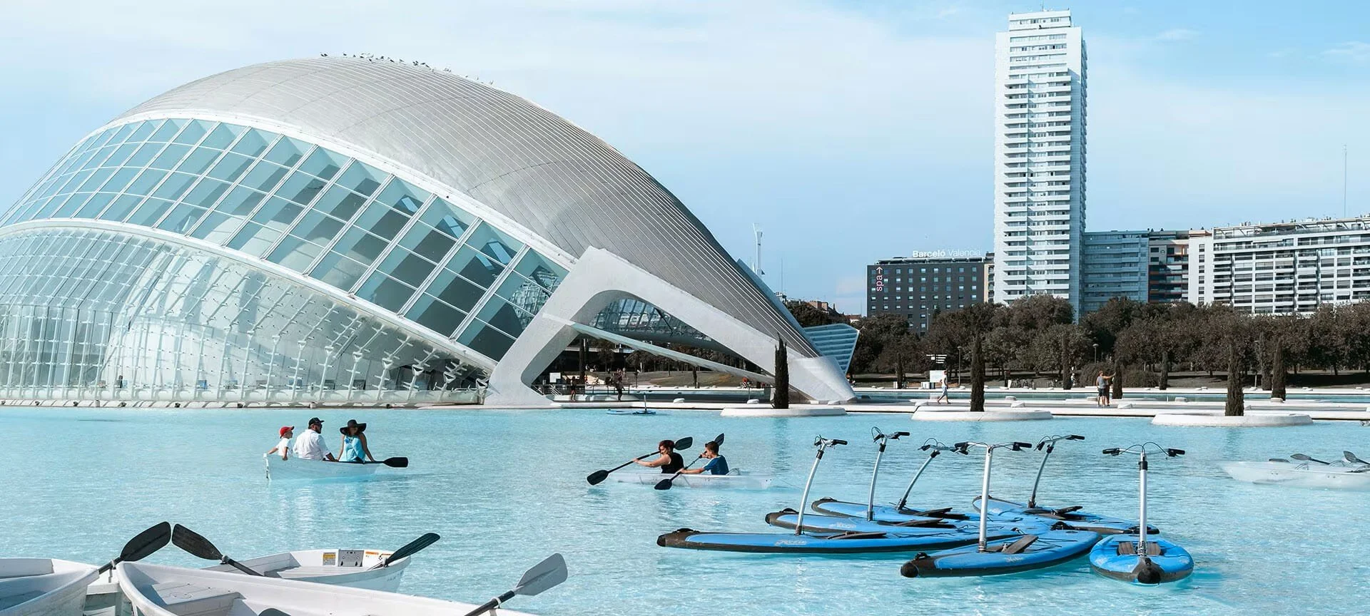 Landscape view of the City of Arts and Sciences complex in Valencia, Spain, featuring a modern, curved building with glass and metallic surfaces, a water feature with paddle boats and people kayaking, trees lining the area, and tall buildings in the background under a blue sky.