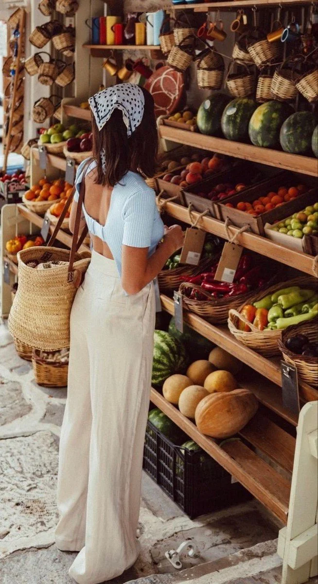 A woman shopping for fruits and vegetables at a produce store, wearing a light blue top and cream-colored wide-leg pants, with a wicker basket purse on her shoulder, and a blue polka dot scarf on her head.