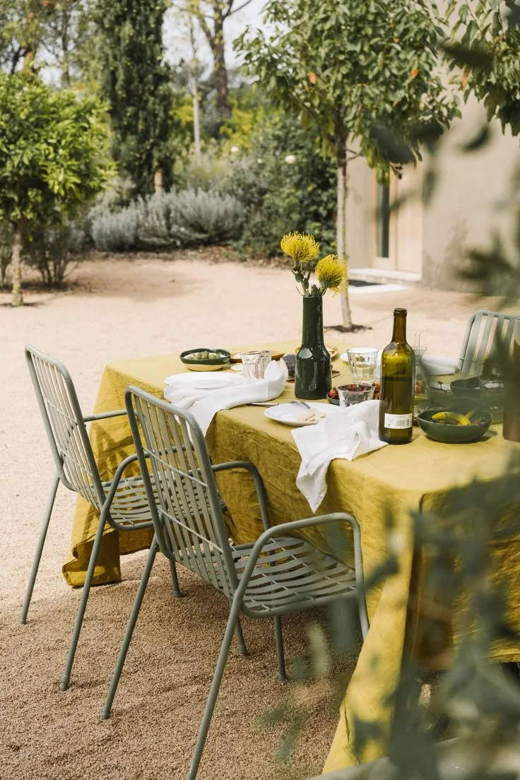Outdoor dining table with yellow tablecloth, surrounded by four metal chairs, set with a black vase containing yellow flowers, bottles, bowls, glasses, and napkins, in a garden with trees and plants in the background.