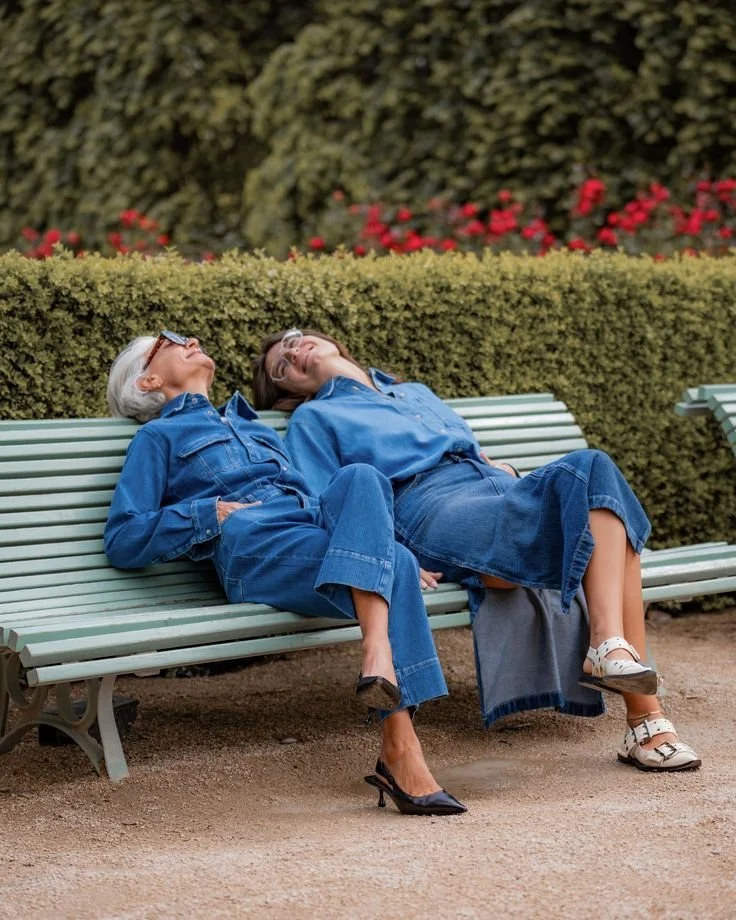 Two older women are sitting on a park bench, leaning back and relaxing with their heads tilted up. They are both smiling and enjoying the sunny day. They are dressed in denim outfits, and there are bushes with red flowers behind them.