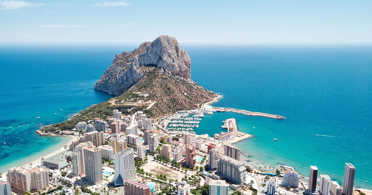 Aerial view of a coastal city with high-rise buildings, a marina, a beach, and a large rocky mountain.
