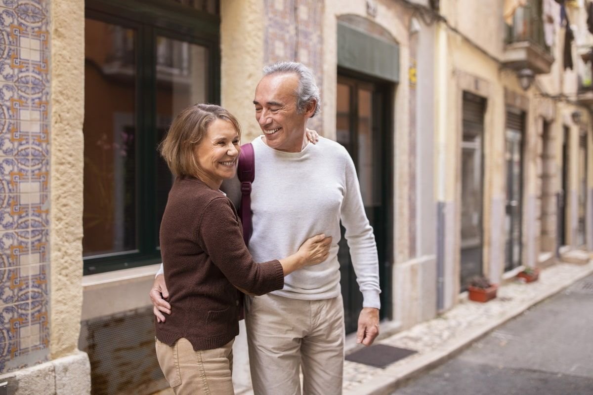 A smiling middle-aged couple embraces on a European street, with narrow cobblestone road and historic buildings with large windows and decorative tiles.