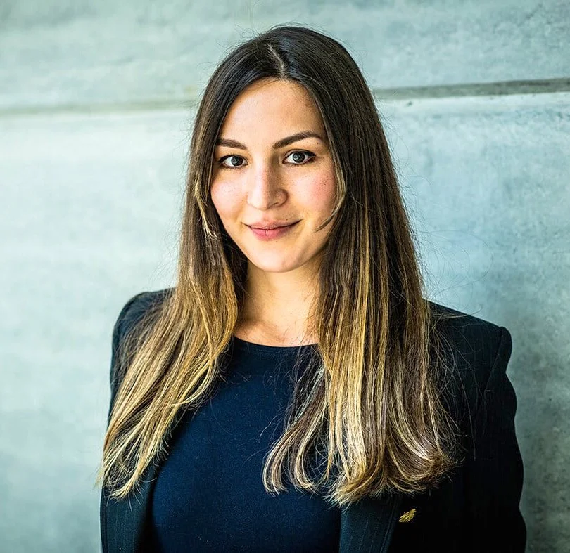 Portrait of a young woman with long brown hair, wearing a black blazer and top, standing against a concrete wall.