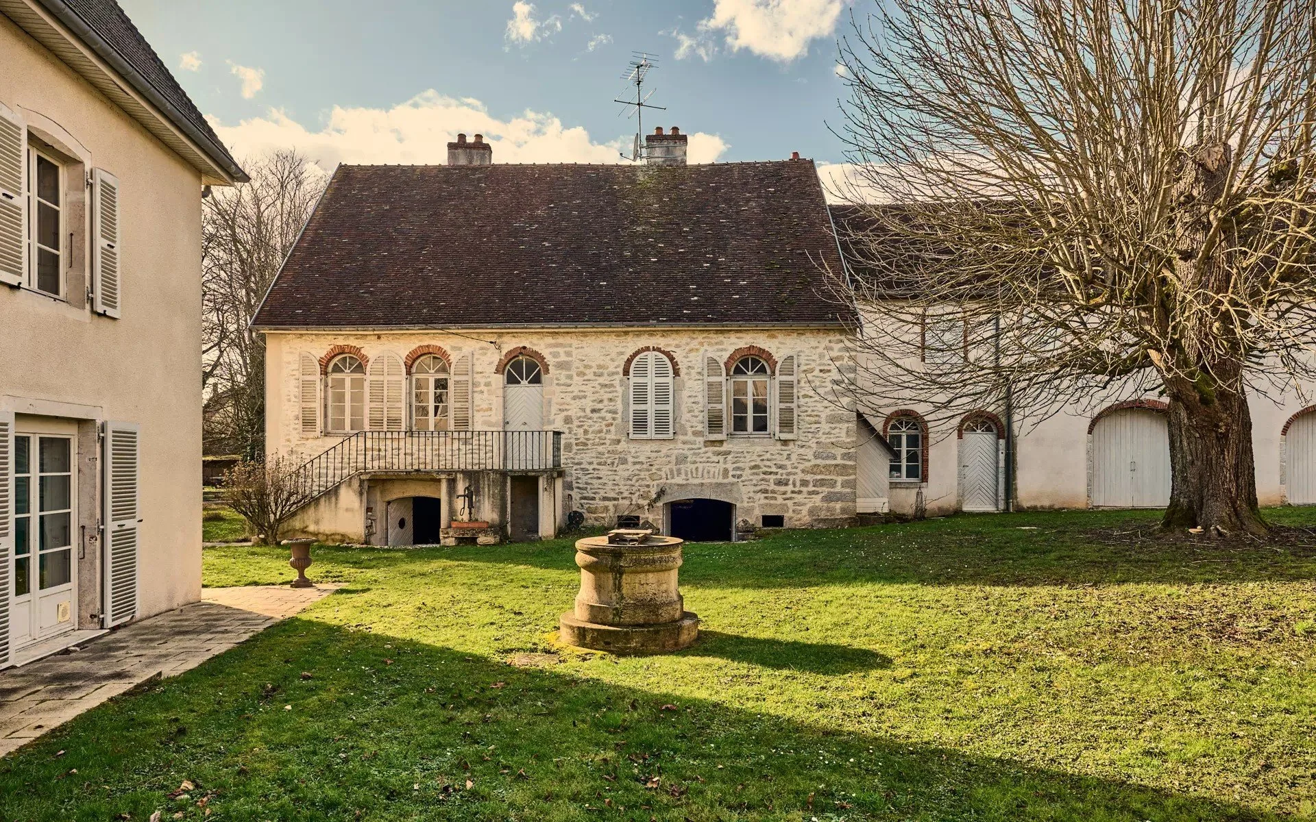 A stone house with a red-tiled roof and white shutters in a yard with grass and a leafless tree.
