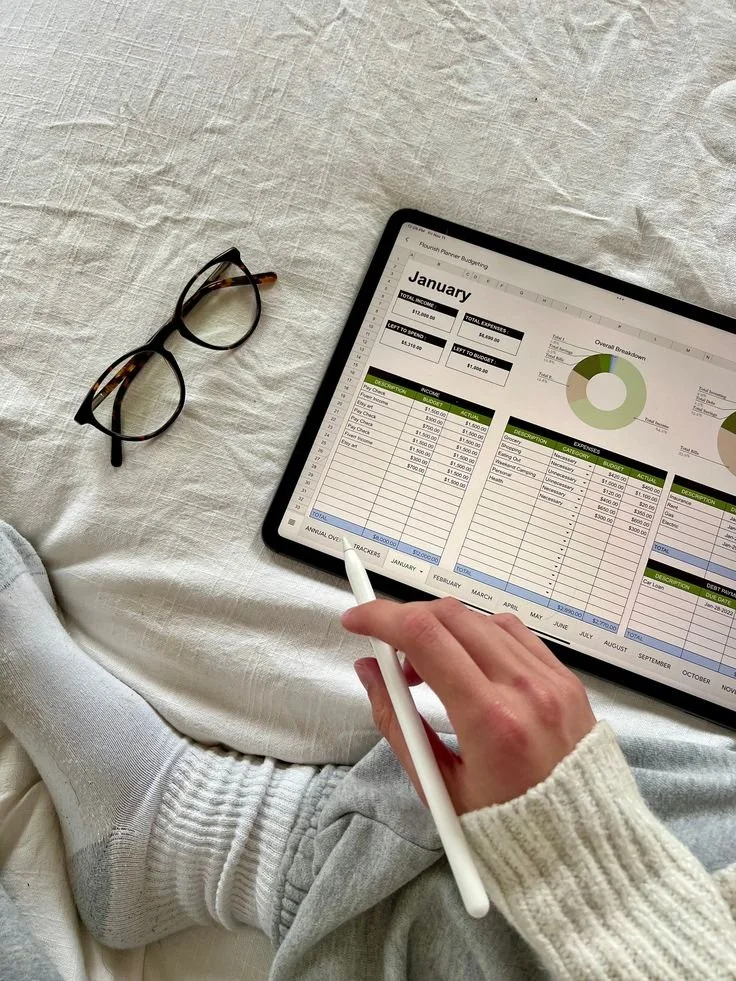 A person sitting on a bed, using a tablet to view a financial budget spreadsheet for January, with a pair of glasses nearby.