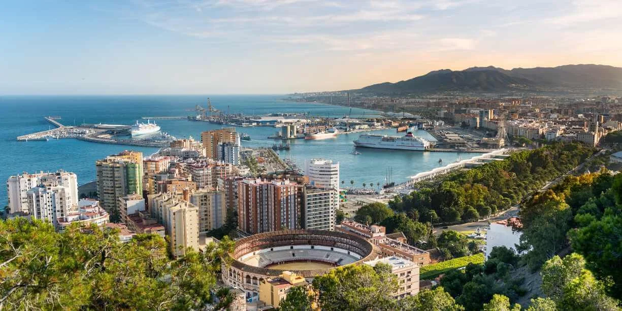 Aerial view of a coastal city with a harbor, high-rise buildings, a stadium, and green hills in the background.
