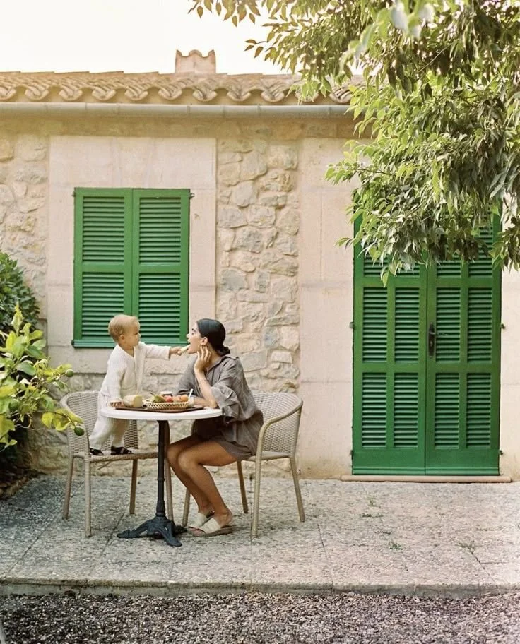 A woman and a young boy sitting at an outdoor table with green shutters and stone wall in the background. The boy is reaching out to the woman's face.