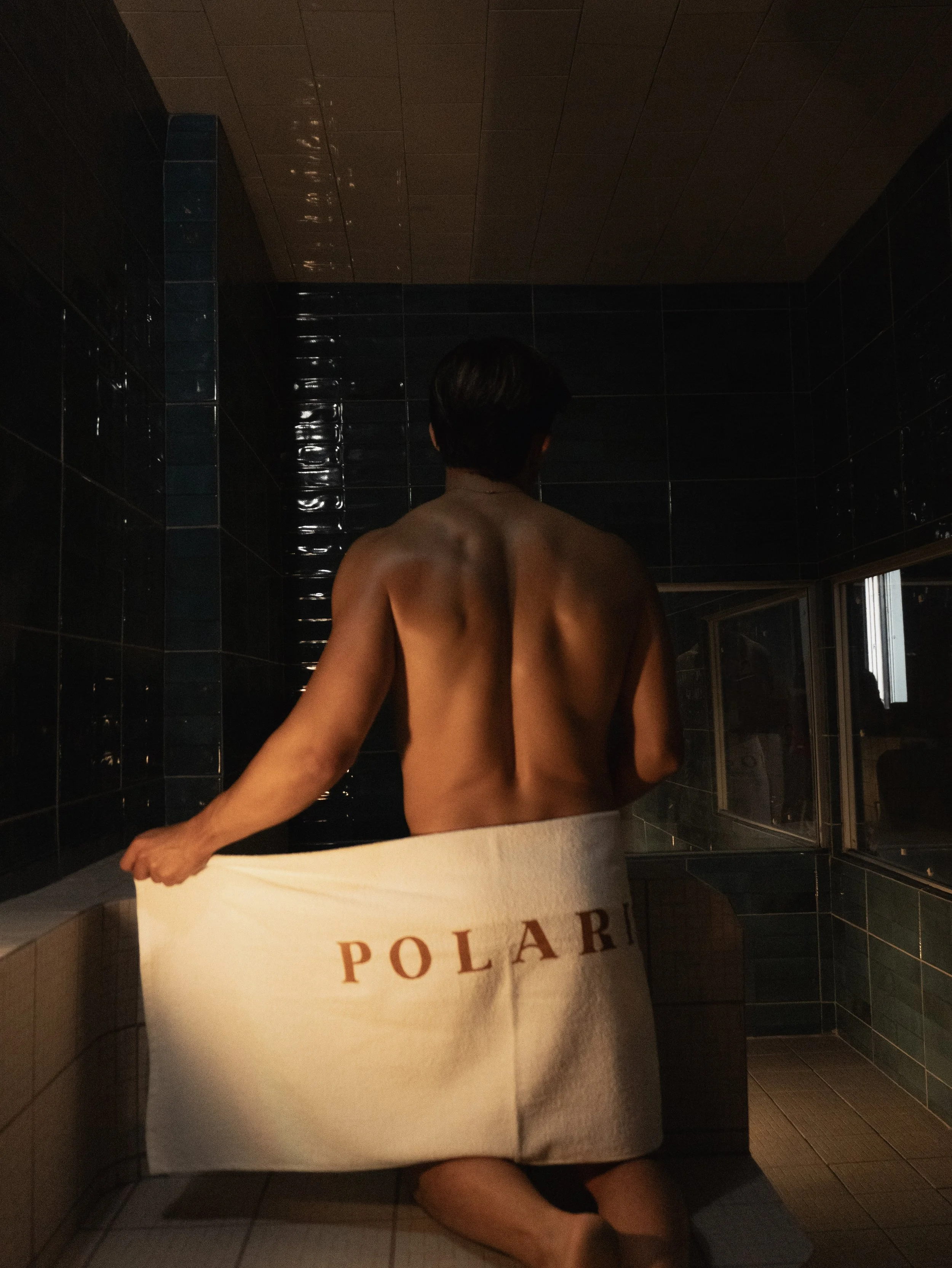Una persona arrodillada en una ducha o baño de vapor, envuelta en una toalla con que dice "POLARI", que es el mejor sauna gay de Guadalajara, de espaldas a la cámara y mirando hacia una pequeña ventana.