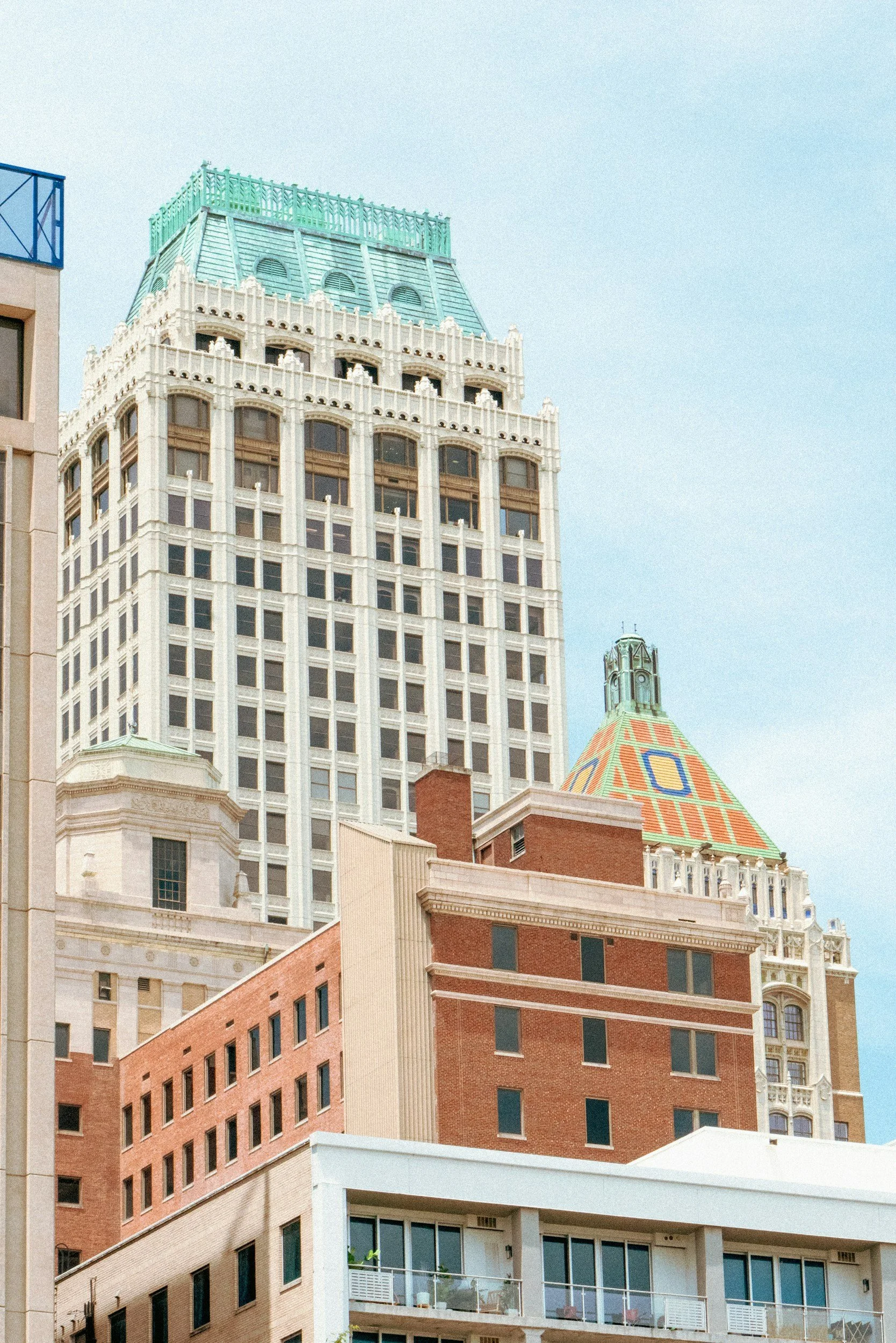 Cityscape with multiple high-rise buildings, including a tall white building with intricate architectural details and a green rooftop, and a shorter building with a colorful, patterned roof, under a clear blue sky.