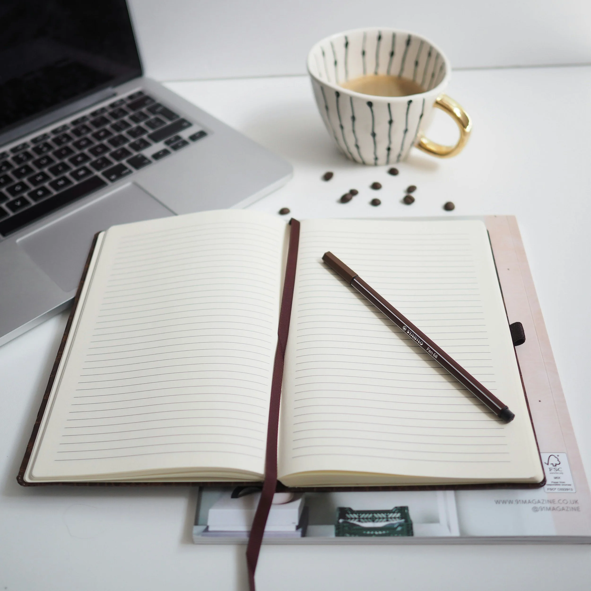 Open notebook, pen on pages, coffee mug, scattered coffee beans, laptop in the background on a white surface.