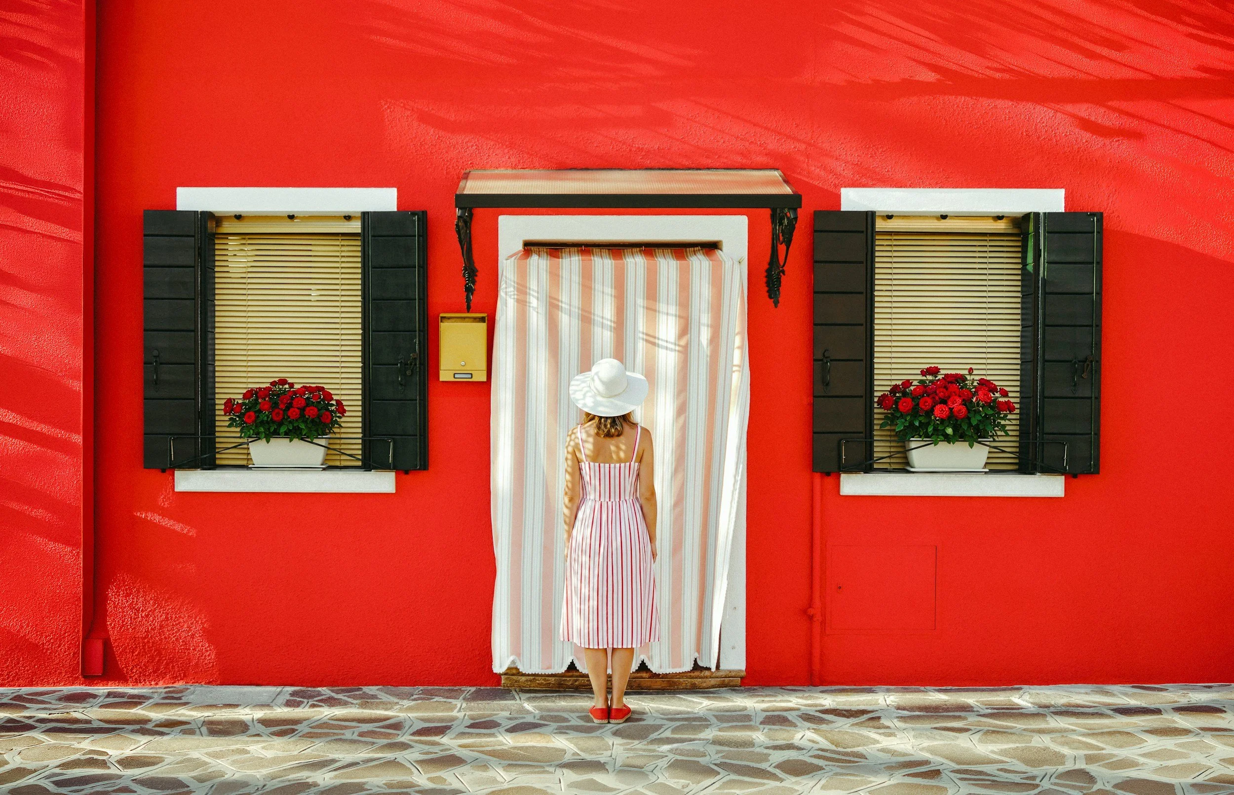 A woman in a striped dress and white sun hat stands outside a red building, facing a window with black shutters and flower boxes with red flowers. She appears to be looking at the window or preparing to enter.
