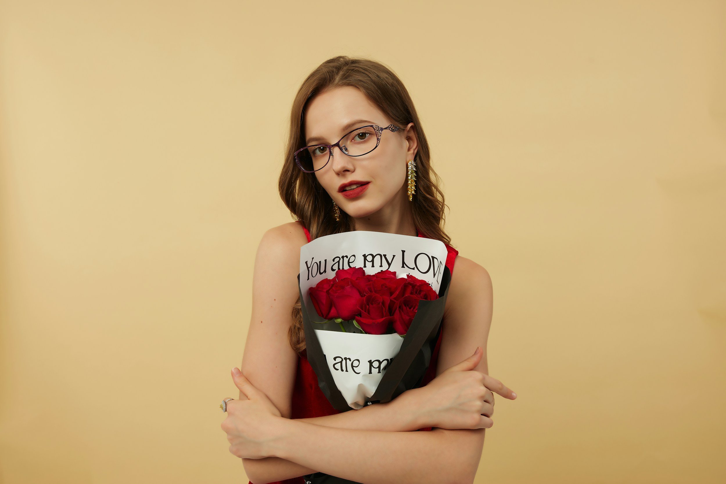 Smiling woman with wavy brown hair and glasses holding a bouquet of red roses wrapped in paper that reads "You are my love."