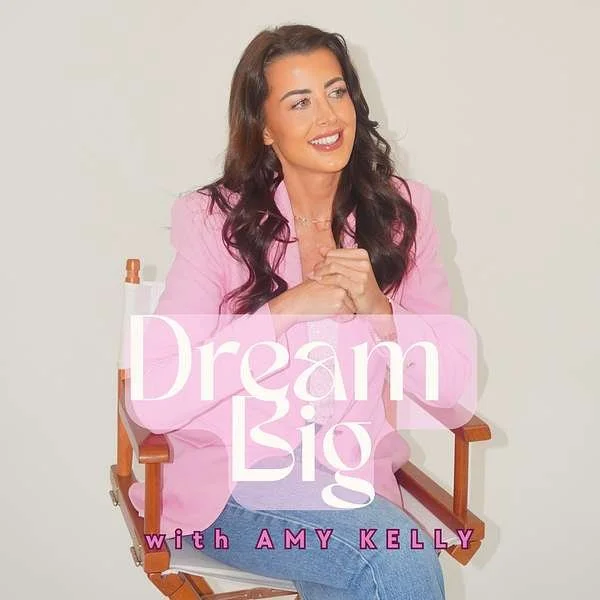 A woman with long dark hair, wearing a pink blazer, sitting on a director's chair. The text on the image says "Dream Big with Amy Kelly."
