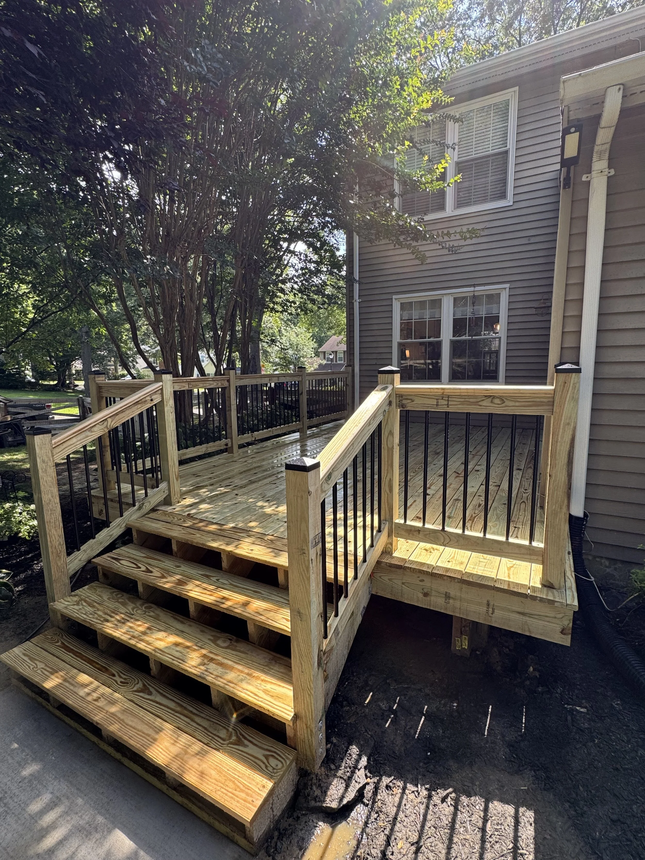 Newly built wooden deck with stairs and black metal railings attached to a beige house with a second story window. Trees and neighboring houses in the background.