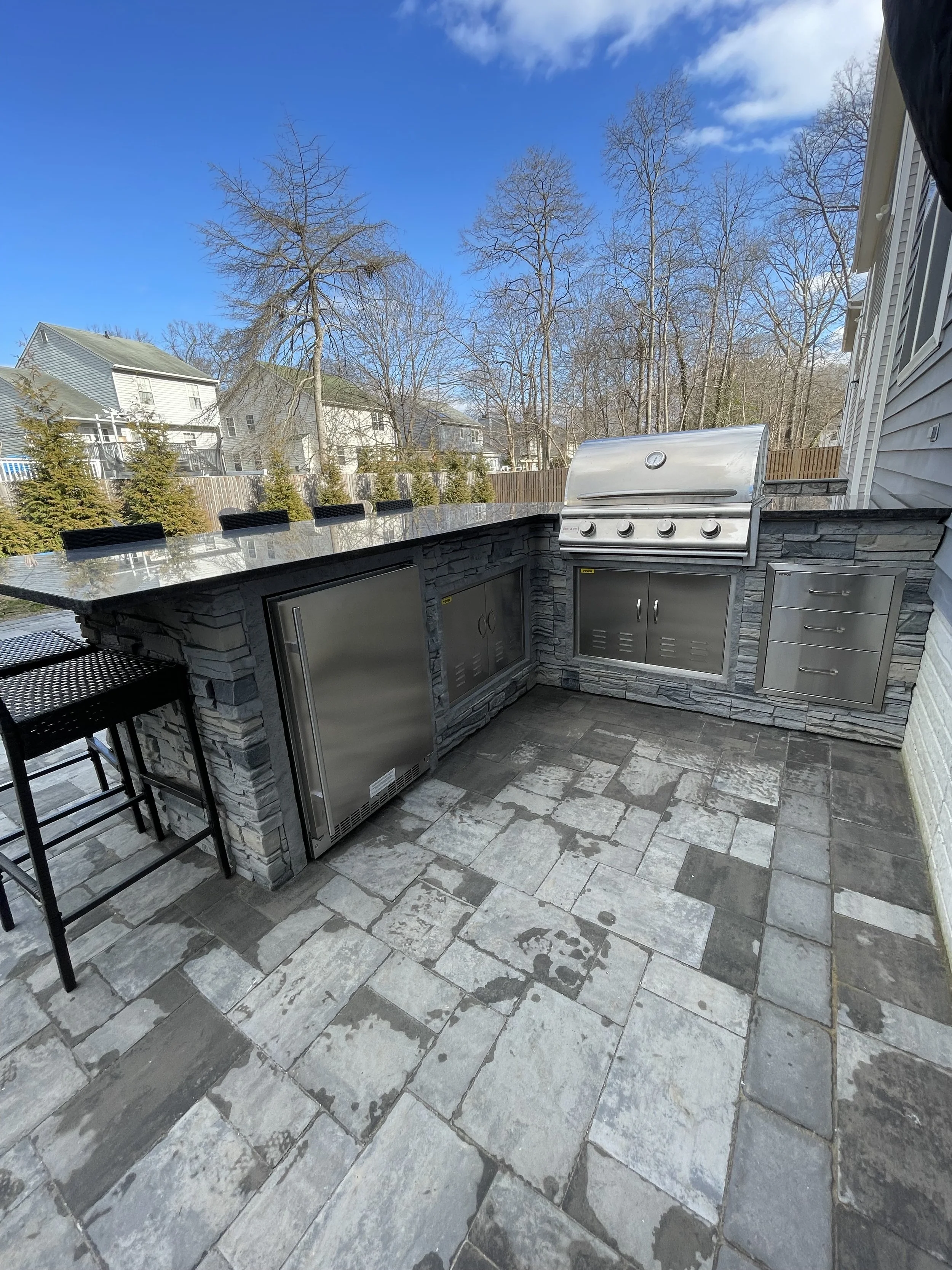 Outdoor kitchen with gray stone cabinets, stainless steel grill, stone backsplash, and bar seating overlooking a backyard with trees and houses under a blue sky.