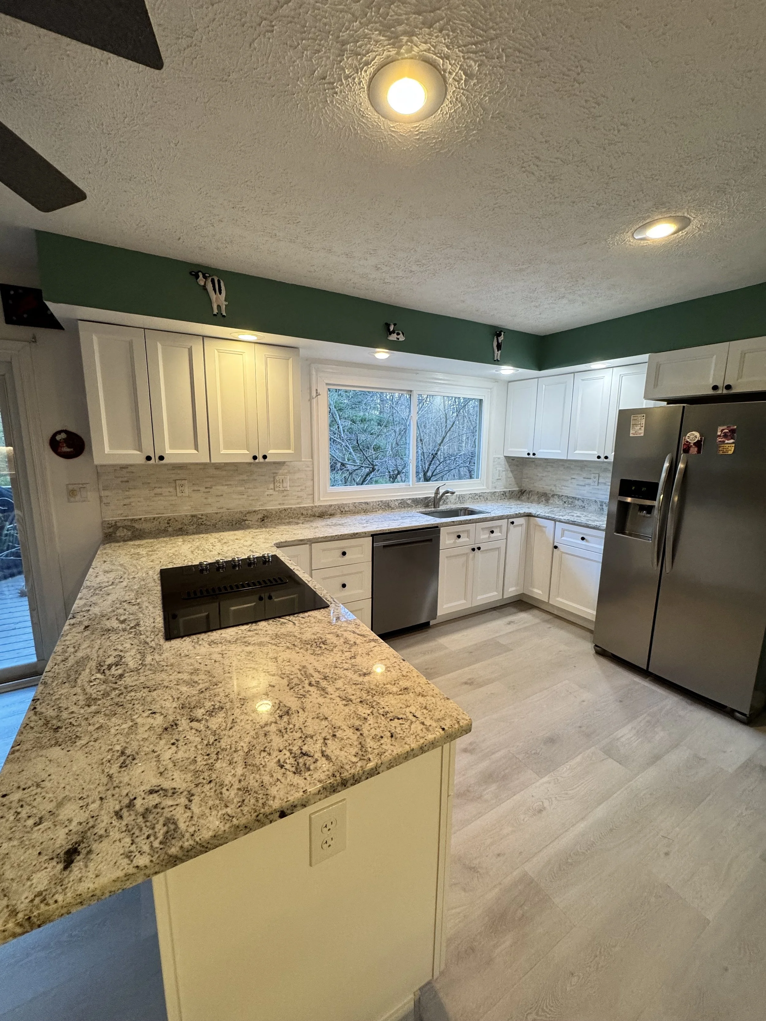 A modern kitchen with white cabinets, granite countertops, a stainless steel refrigerator, a dishwasher, a stovetop, and a window overlooking a wooded area. The ceiling has recessed lighting and small decorative cow figurines along a green upper wall.