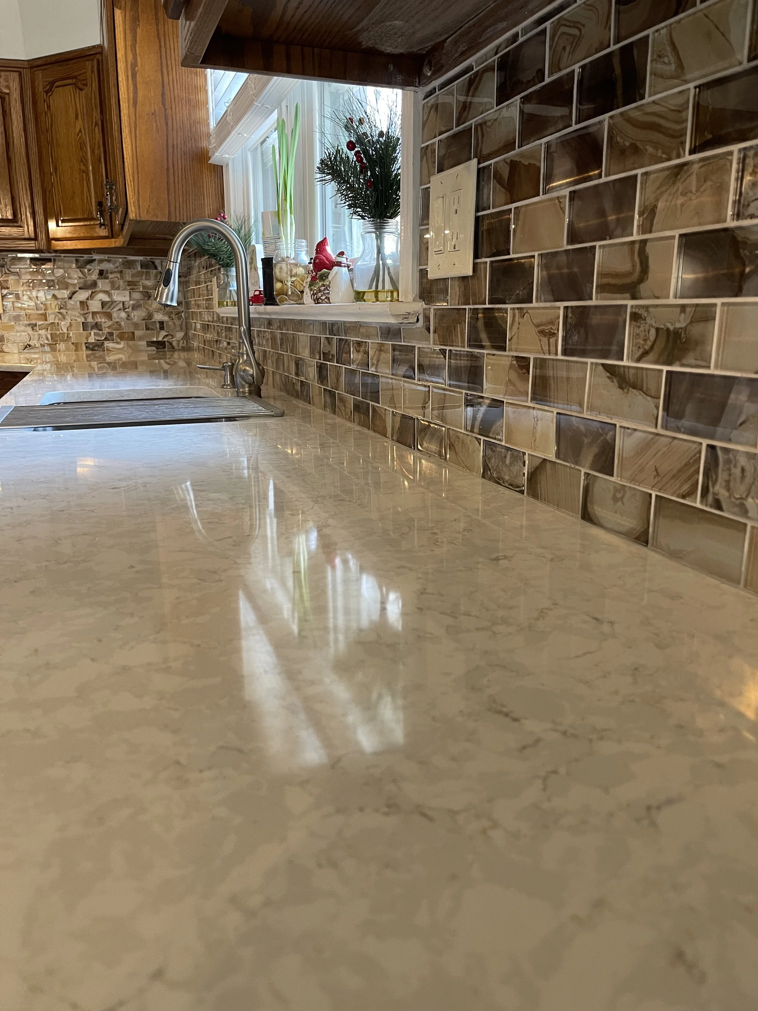 Kitchen countertop with a marble surface, a stainless steel sink with a curved faucet, a window with holiday decorations including a small Christmas tree and a potted plant, and a beige tile backsplash.