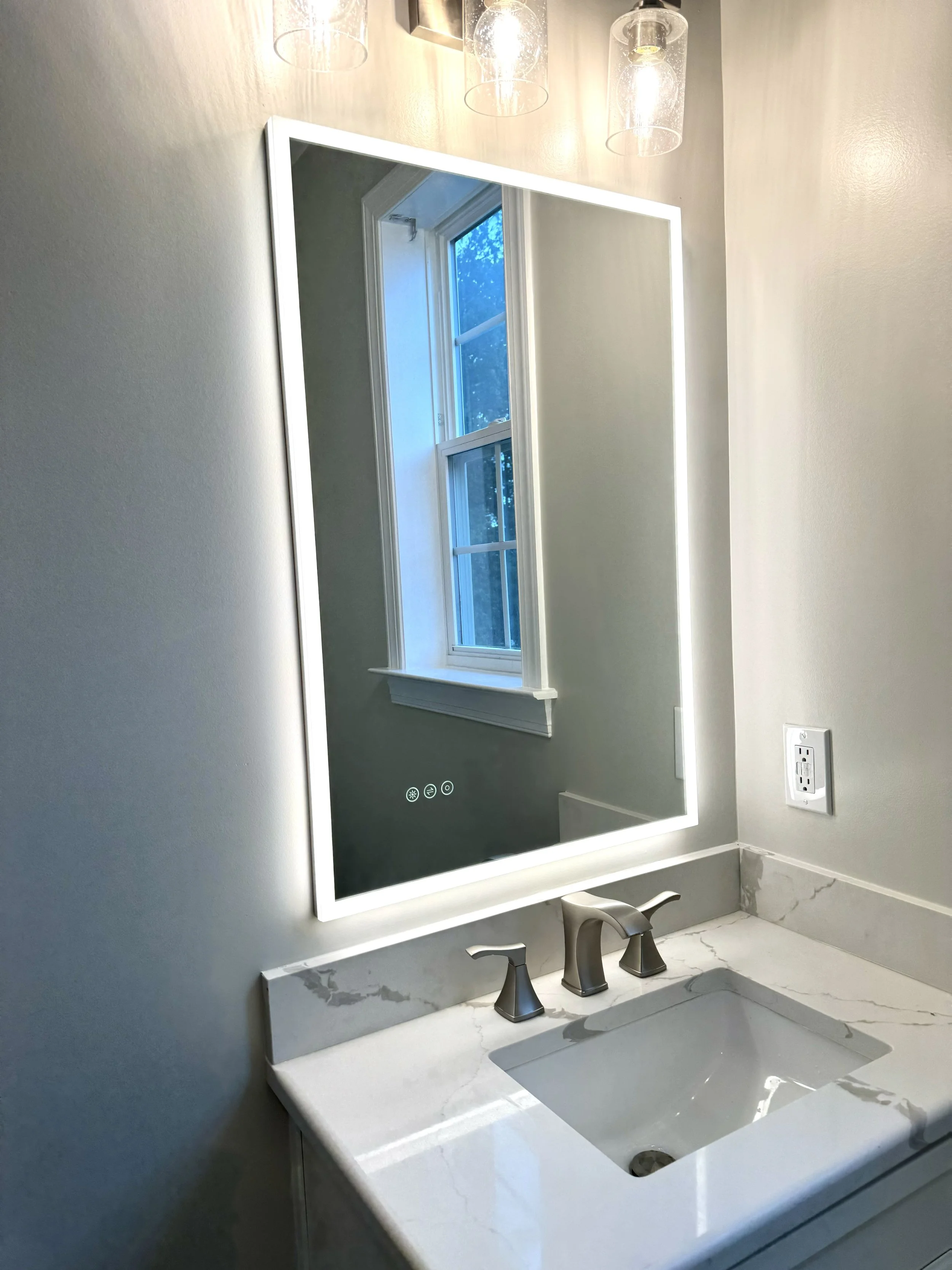 Bathroom vanity with a white marble countertop, undermount sink, and a backlit mirror above it. There is a window with white trim reflecting in the mirror and three pendant lights hanging above.
