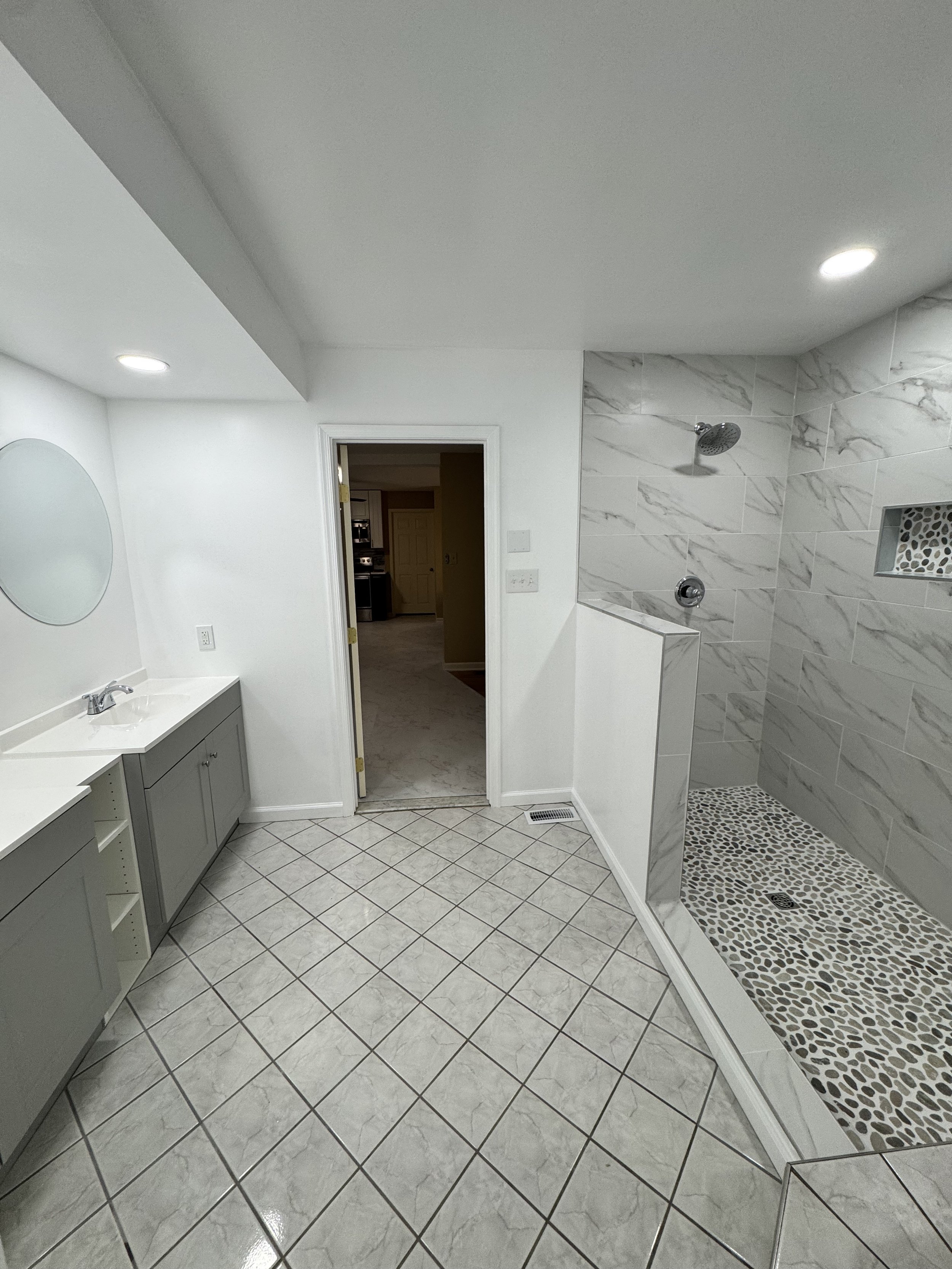 A bathroom with a white countertop vanity, a round mirror, white walls, a tiled shower area with pebble floor, and a doorway leading to another room.