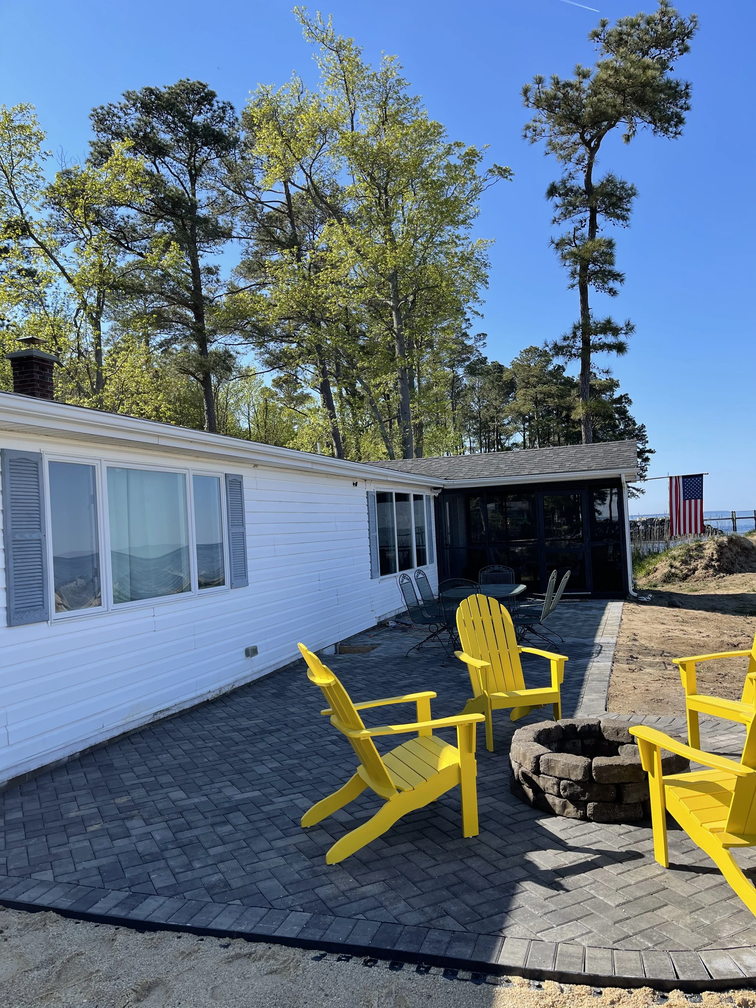 A backyard patio with four yellow Adirondack chairs surrounding a stone fire pit. The patio is paved with dark gray bricks, and there is a white house with blue shutters in the background. Trees are visible behind the house, along with a clear blue sky and an American flag in the distance.