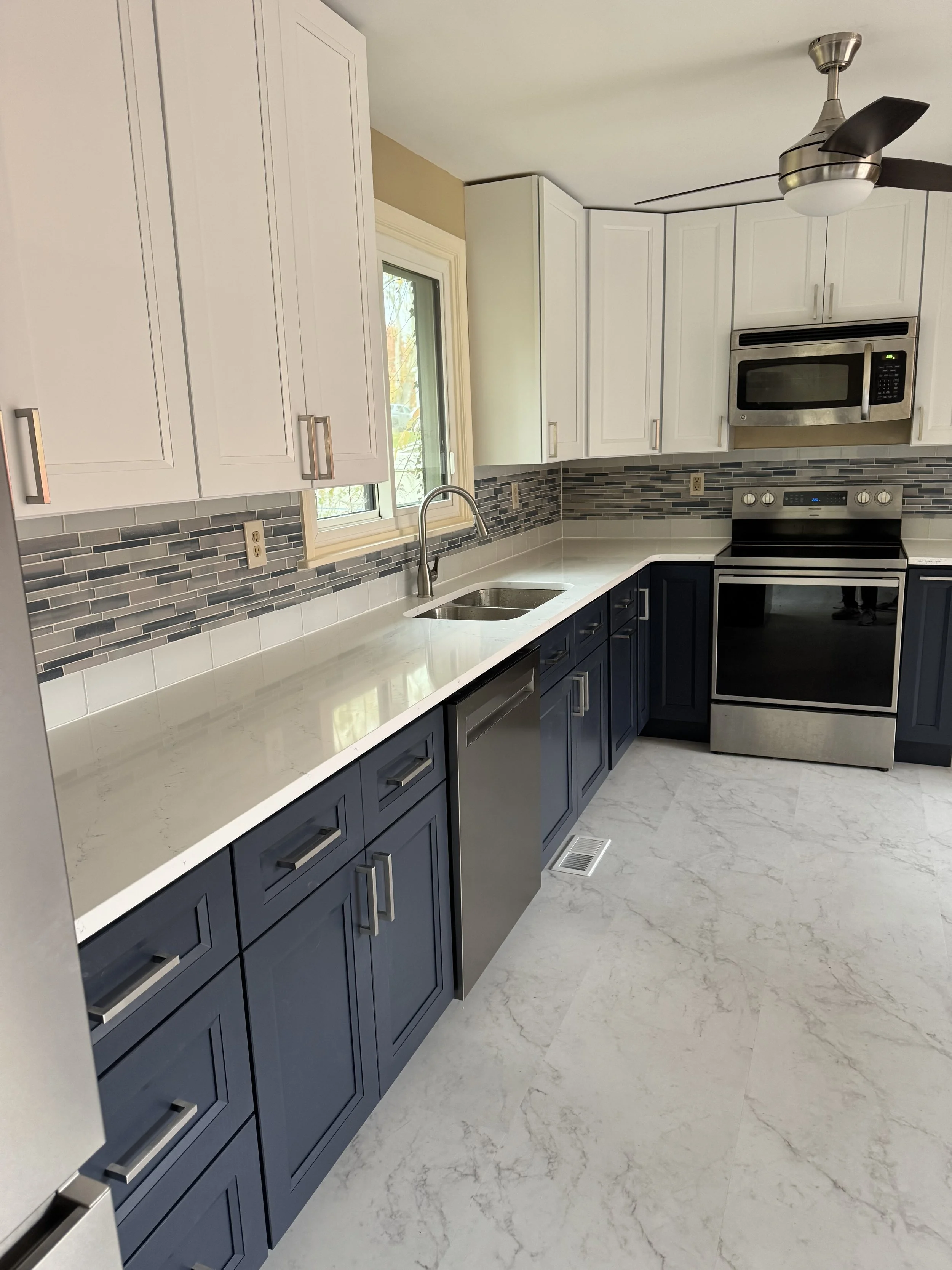 Modern kitchen with white upper cabinets, dark blue lower cabinets, a white countertop, a gray backsplash, stainless steel appliances, and a ceiling fan. There is a window above the sink and a vent on the floor.