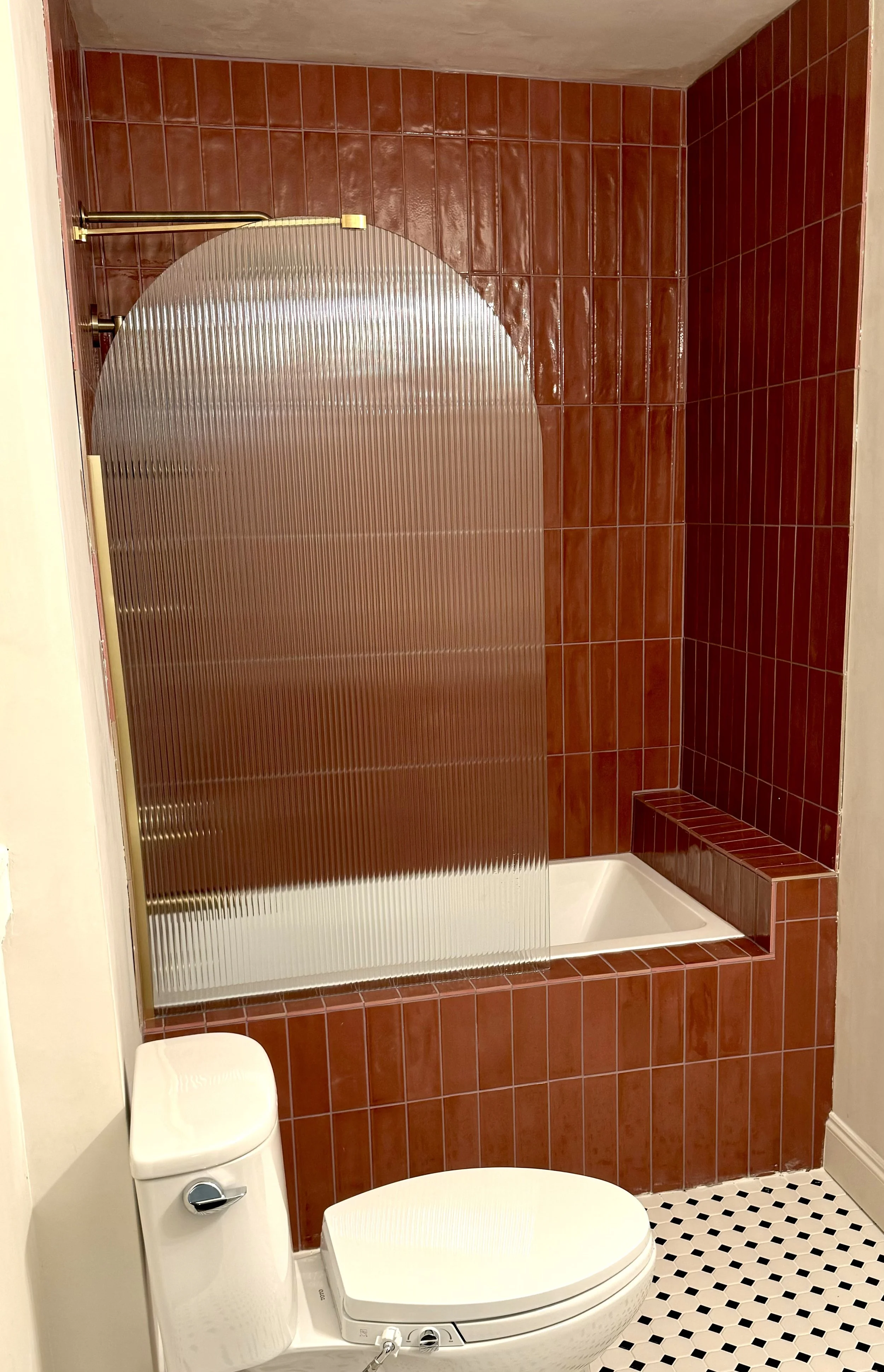 A bathroom with a red tiled wall, a bathtub with a raised edge, a frosted glass shower screen, and a white toilet with a bidet attachment.