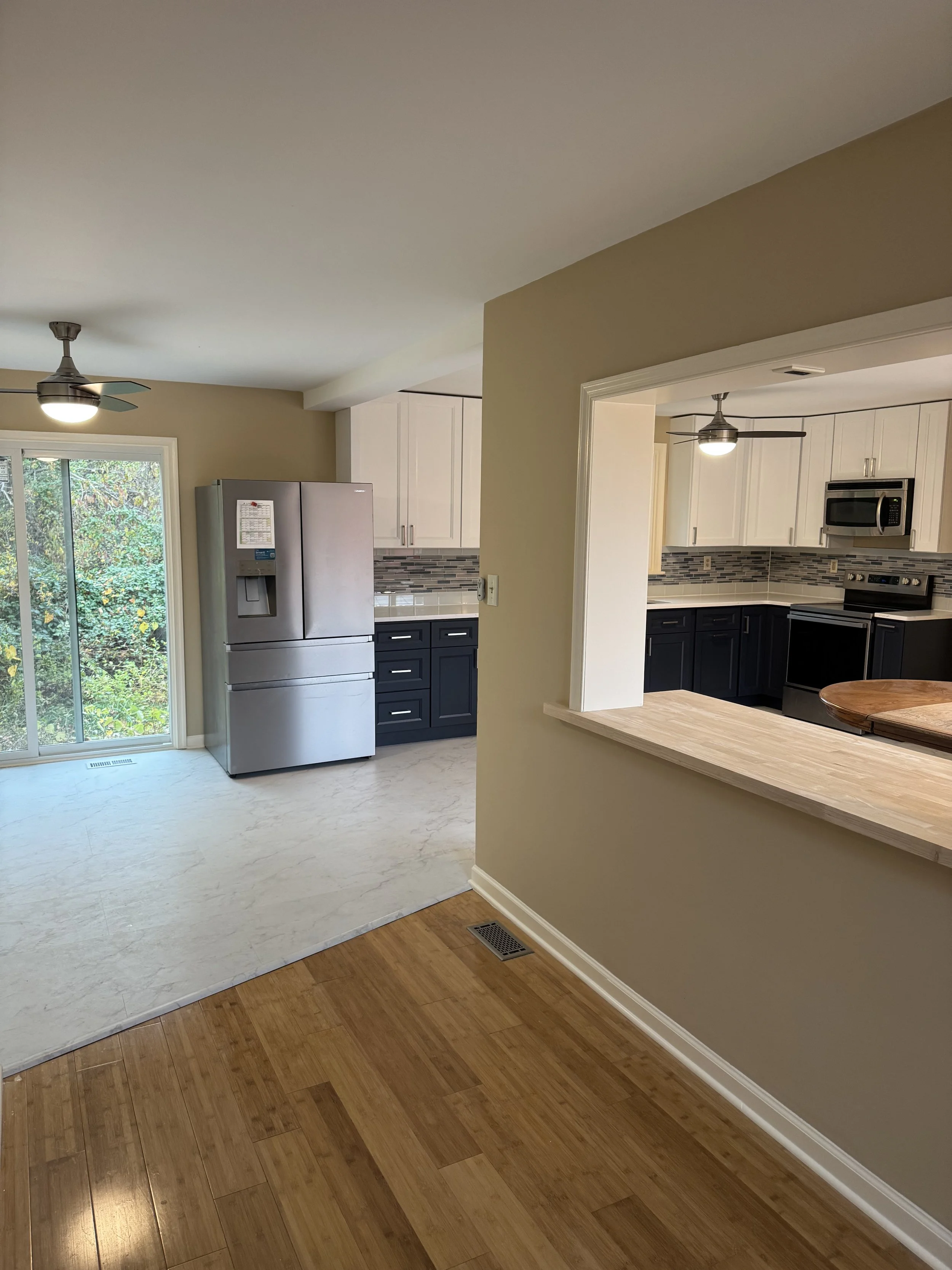 A kitchen with white cabinets, a gray refrigerator, and a colorful tile backsplash. There is a small opening in the wall with a counter extending into the adjoining room which has hardwood flooring. The room features ceiling fans and sliding glass doors with a view of greenery outside.