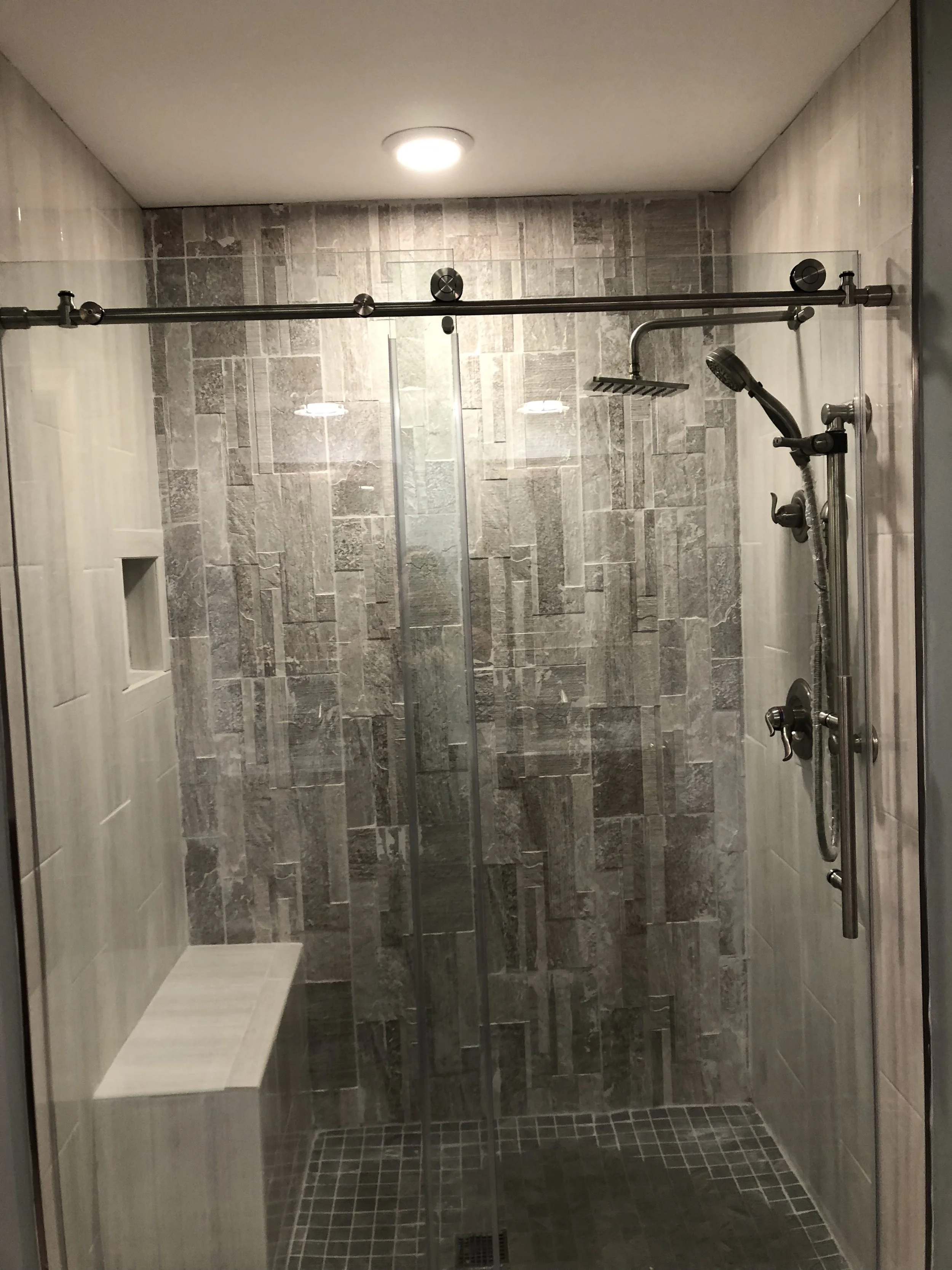 A modern walk-in shower with gray stone tiles, a built-in bench, a glass door, and a rainfall showerhead.
