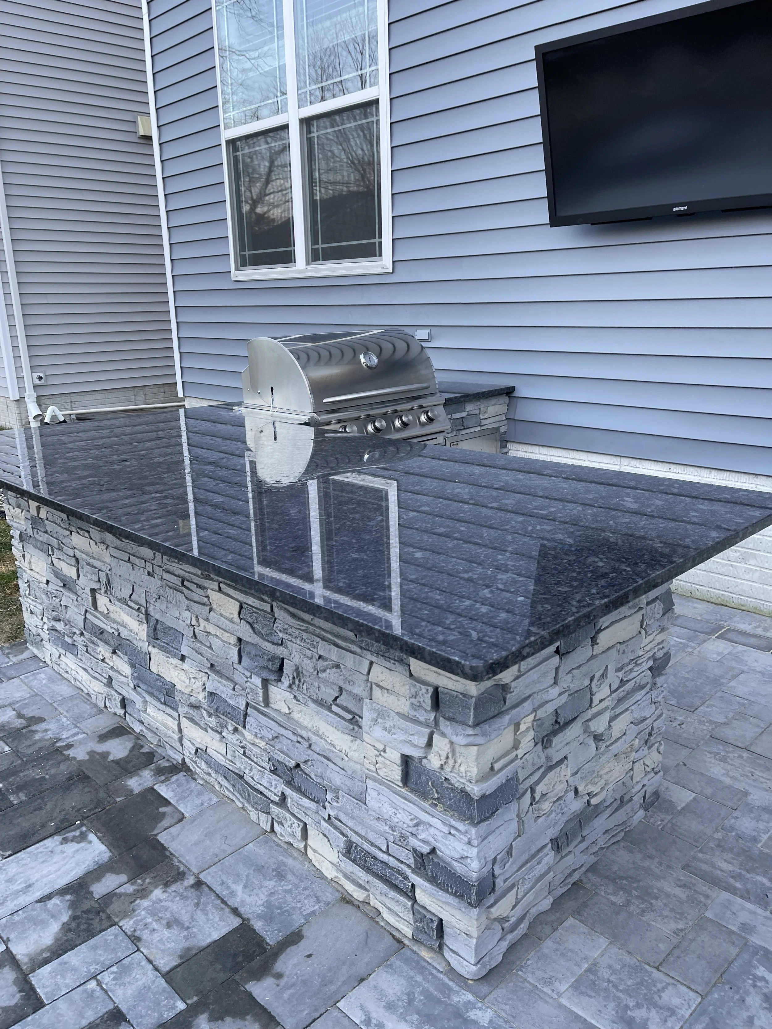 Outdoor kitchen with a black granite countertop, stone base, stainless steel grill, and mounted flat-screen TV on a blue siding house.
