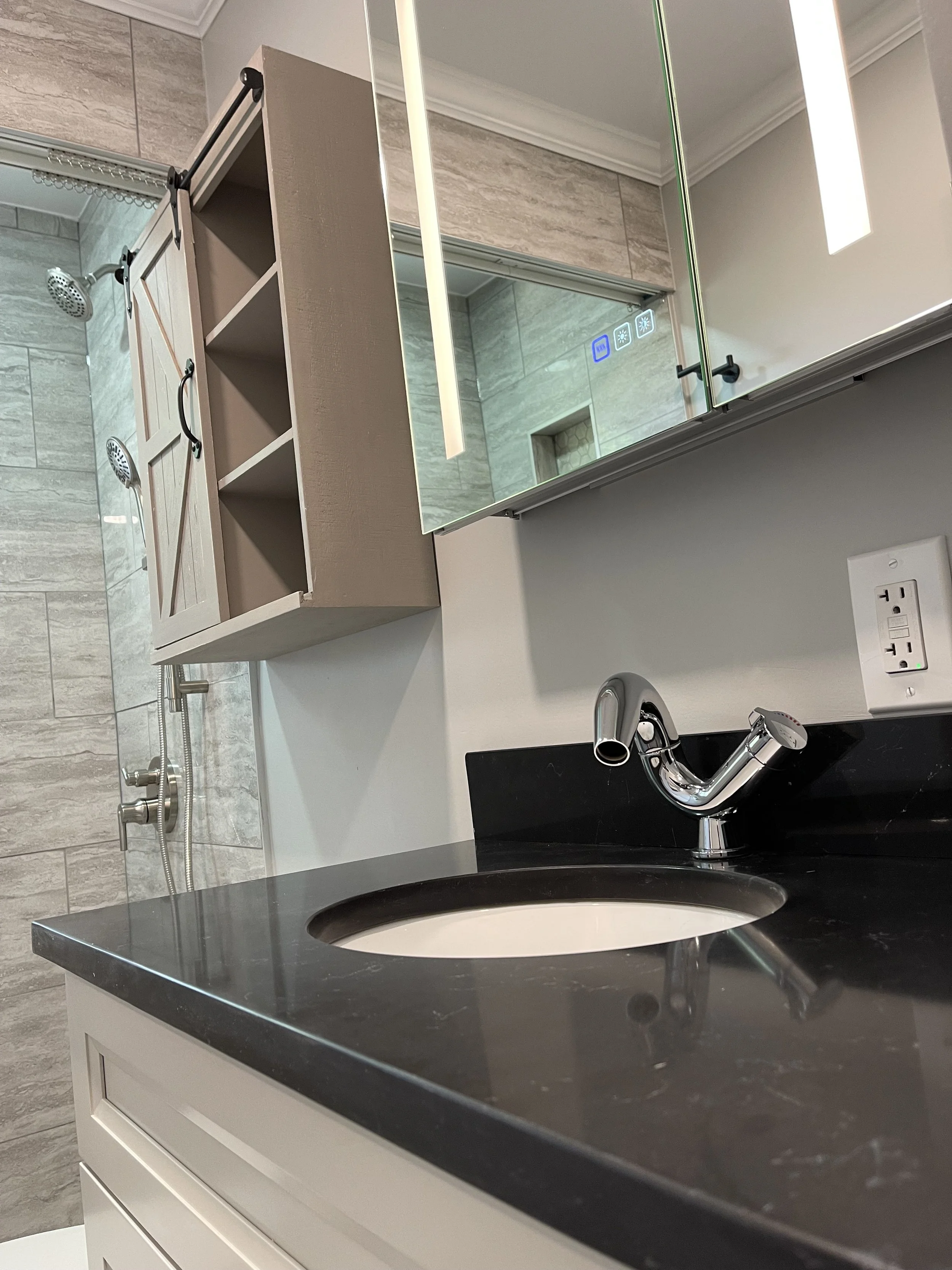 Bathroom vanity with black countertop, oval sink, chrome faucet, white cabinet, mirror, wall outlet, shower area with gray tile and dual showerheads.
