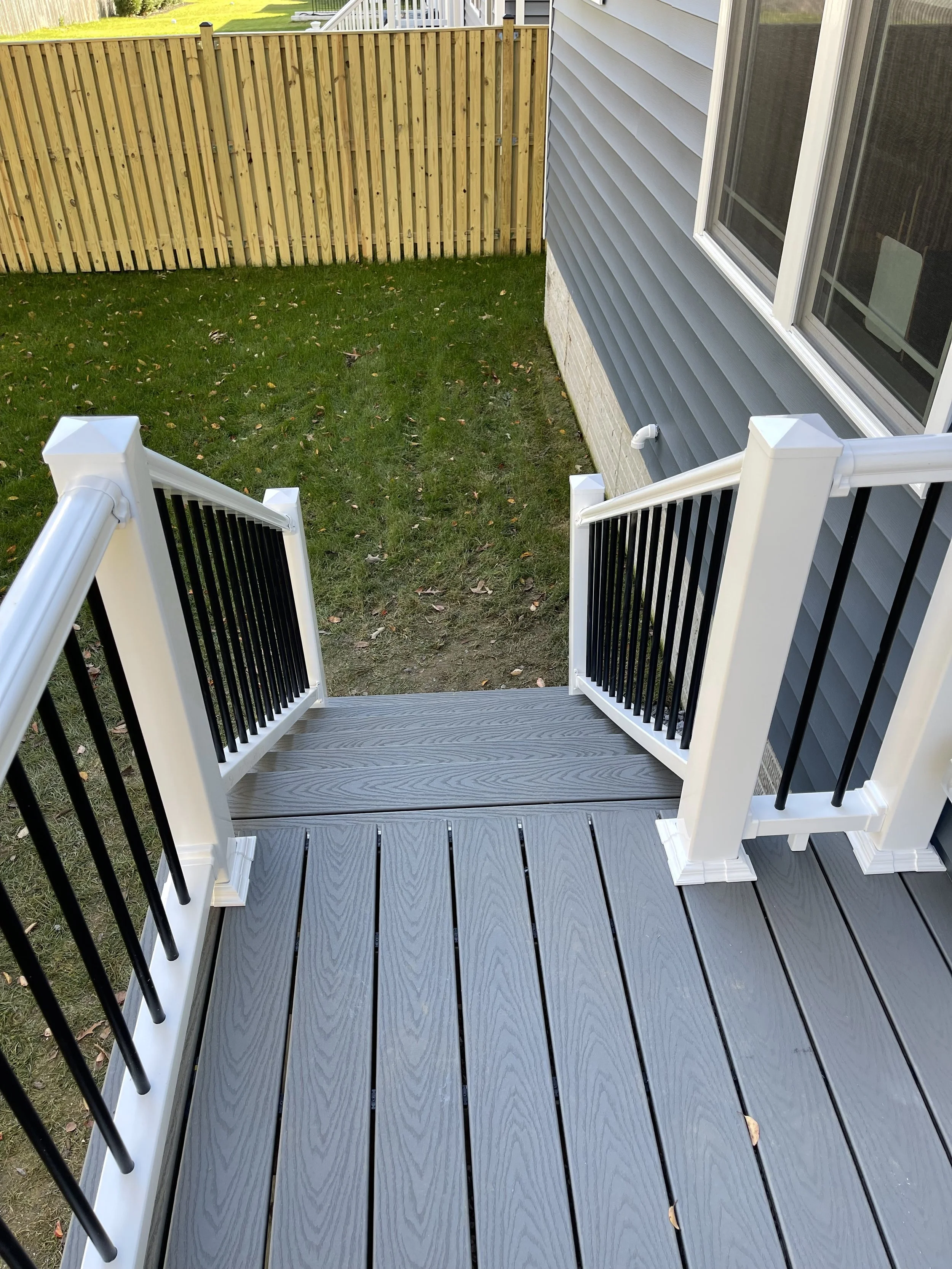 View of a small backyard patio with gray wooden decking, white and black railing, adjacent to a house with blue siding, a window, and a gutter pipe, enclosed by a wooden fence.