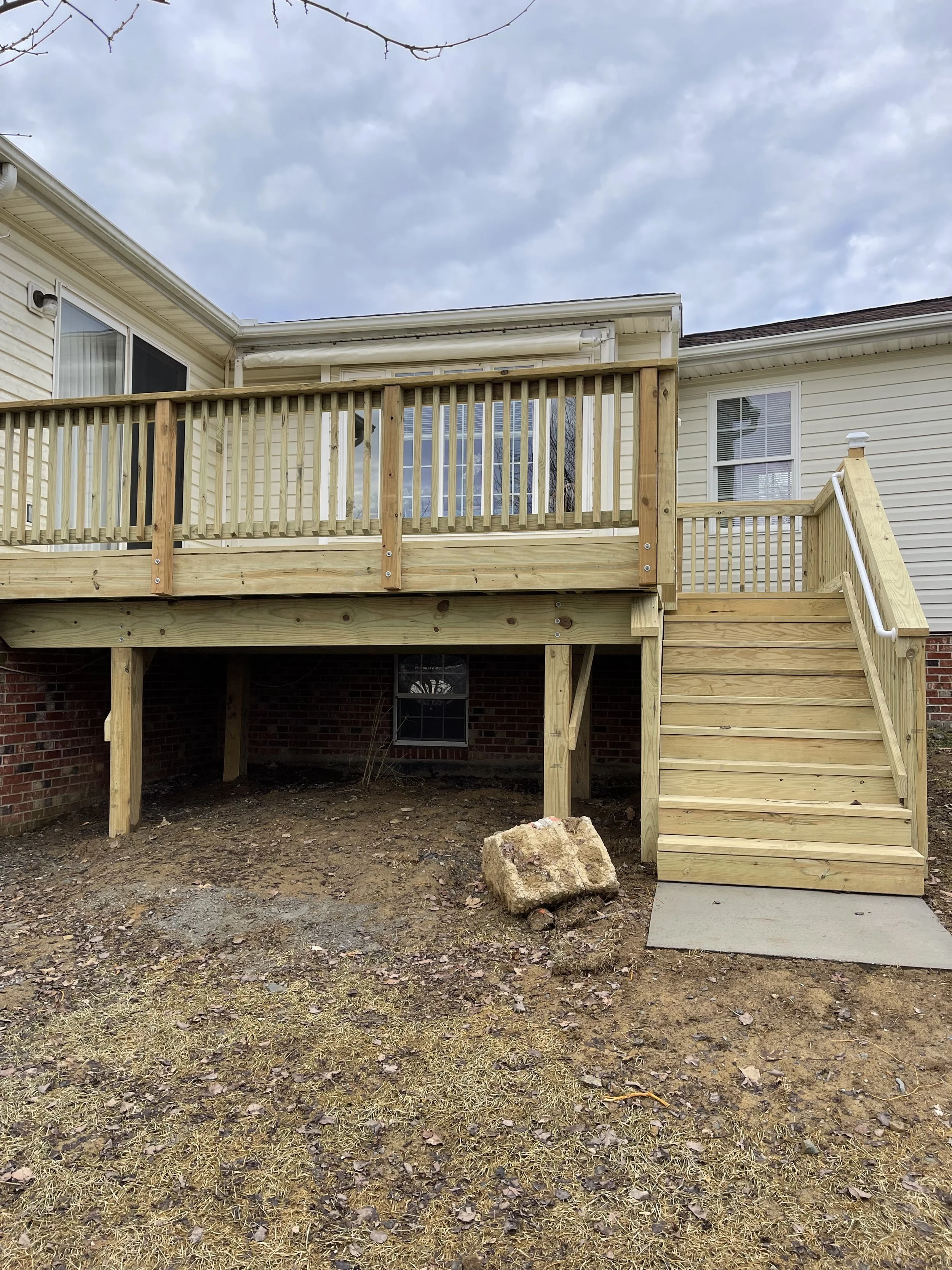 Newly built wooden deck with stairs attached to the back of a house, with dirt and rocks underneath, overcast sky.