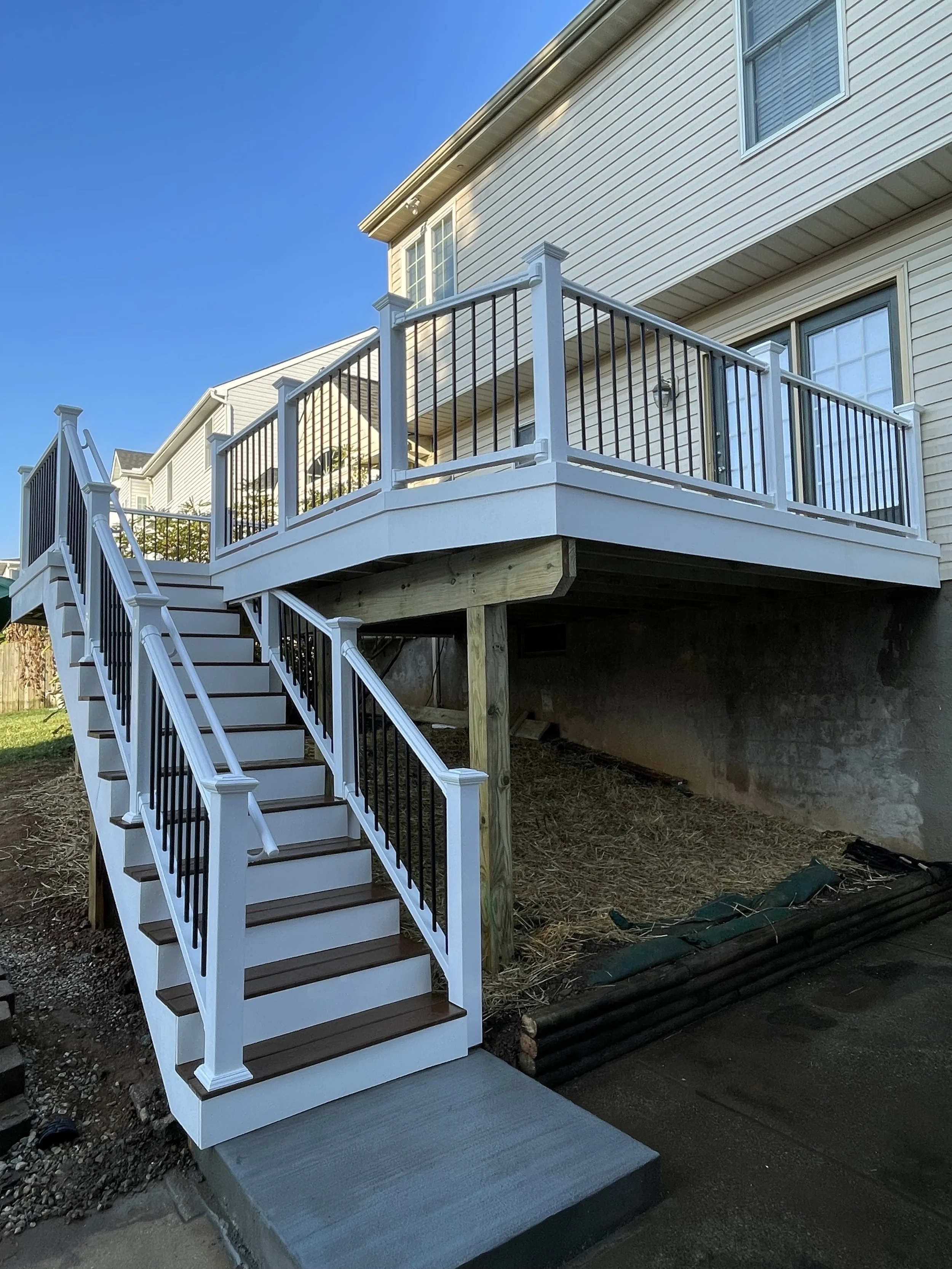 Newly built white deck with black and white railing and stairs attached to beige house, with blue sky background.