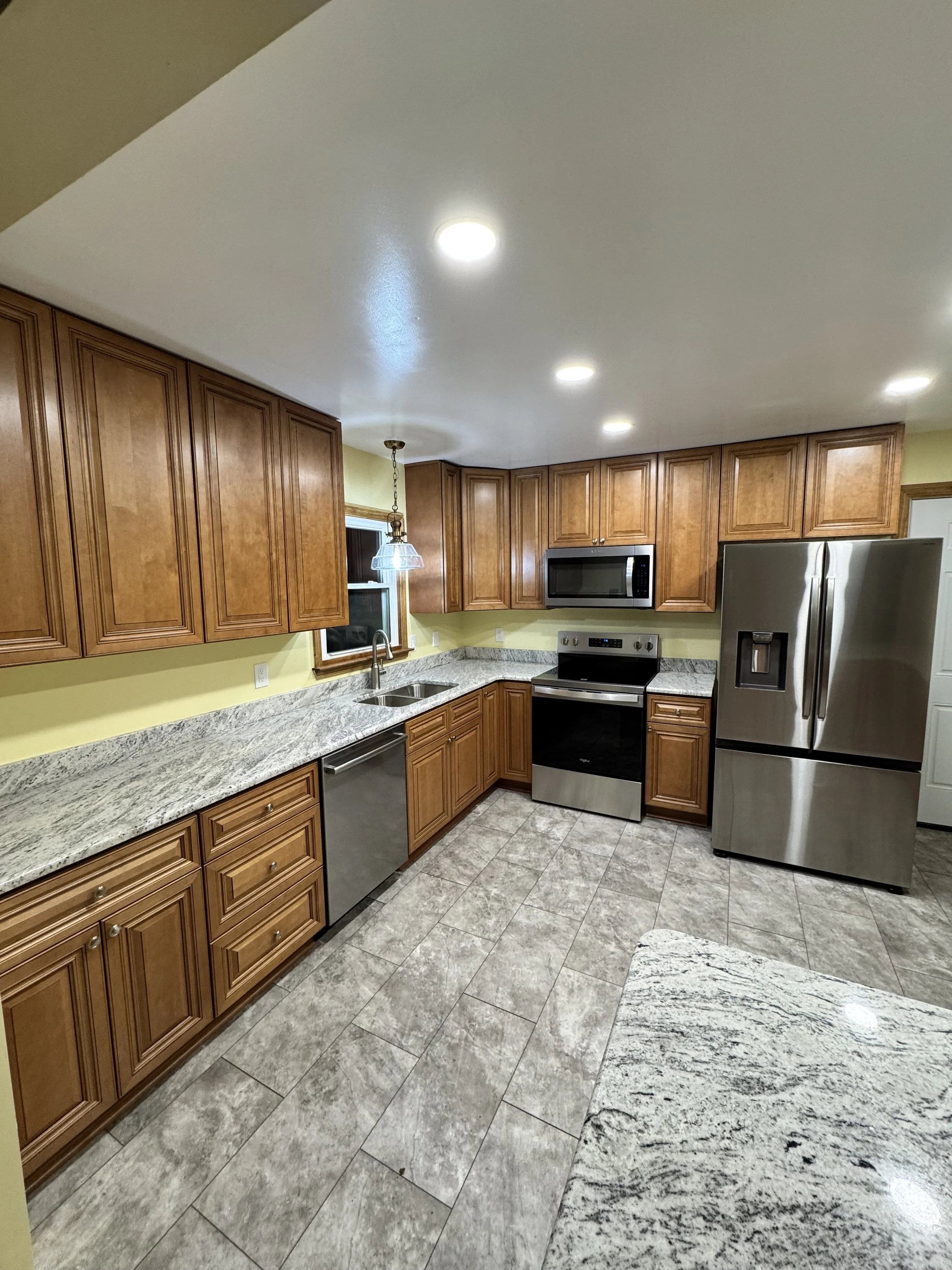 Kitchen with wooden cabinets, granite countertops, stainless steel refrigerator, stove, microwave, dishwasher, tiled floor, and a small window above the sink.