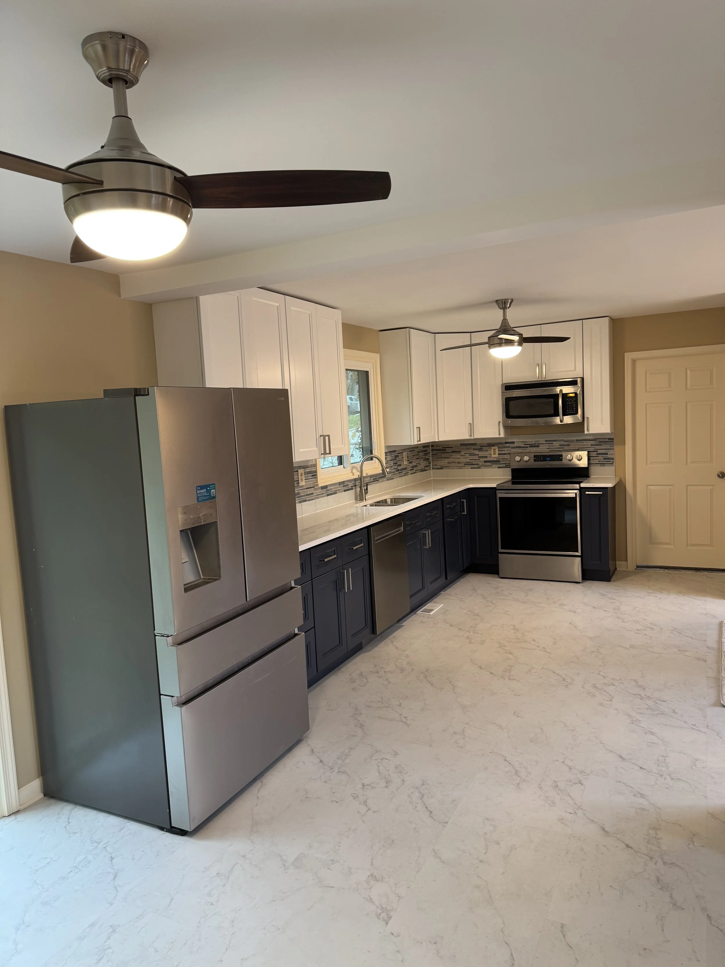 Modern kitchen with a stainless steel refrigerator, white upper cabinets, dark lower cabinets, a built-in microwave, and a stove. Ceiling fans with light fixtures are mounted on the ceiling.