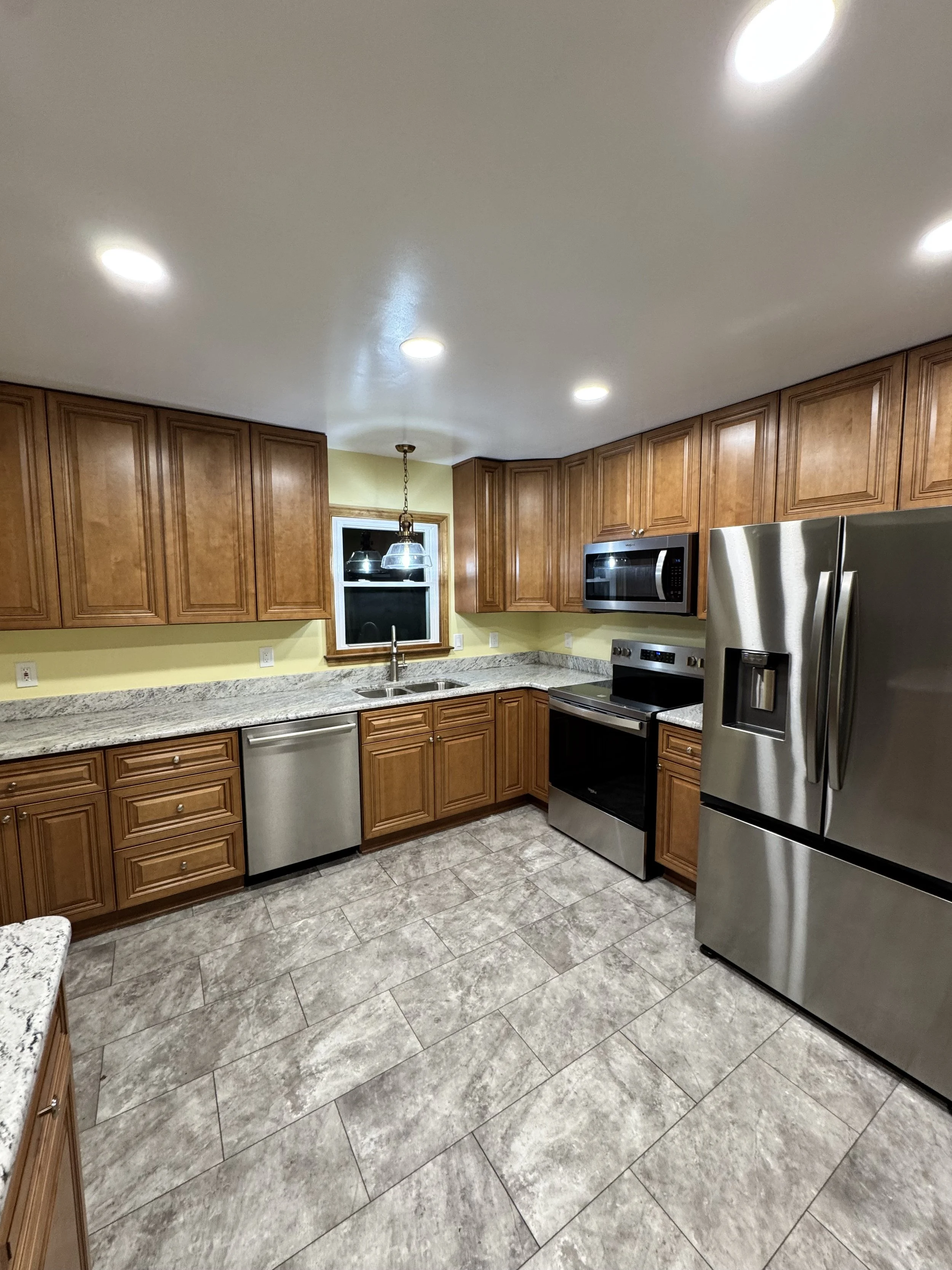 A spacious kitchen with wooden cabinets, granite countertops, stainless steel appliances including a refrigerator, oven, microwave, and dishwasher. There is a small window above the sink and a hanging light fixture.