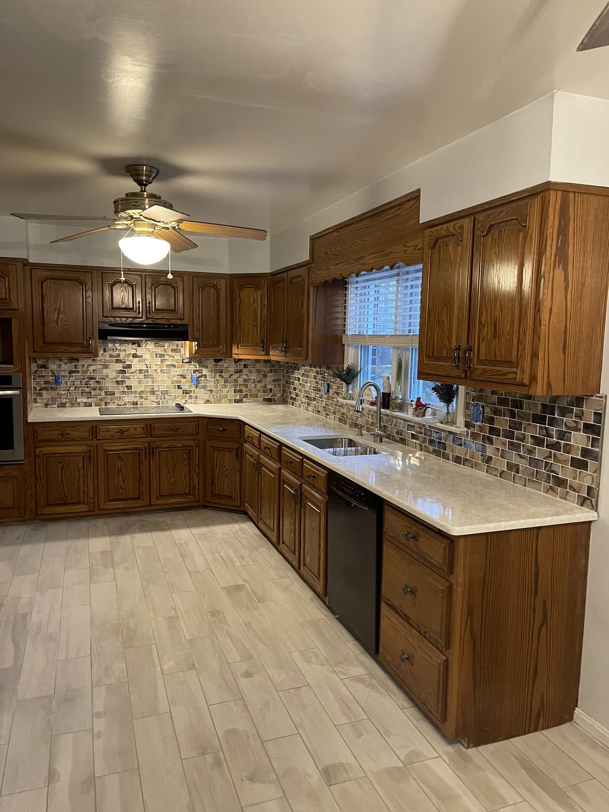 Kitchen with wooden cabinets, tile backsplash, granite countertops, stainless steel sink, dishwasher, and a window with blinds, decorated with small plants and holiday figures.