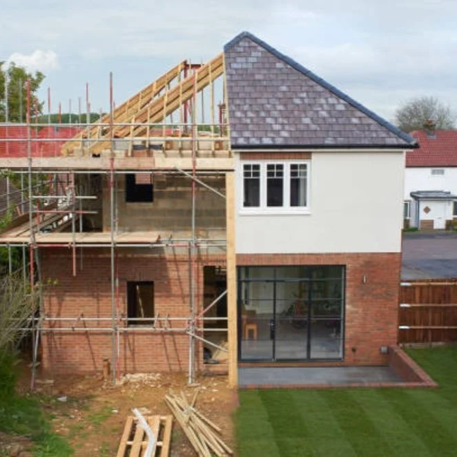 Side view of a two-story house under renovation with scaffolding on the left side, showing brick and white-painted exterior walls, large front window, and a sloped roof.