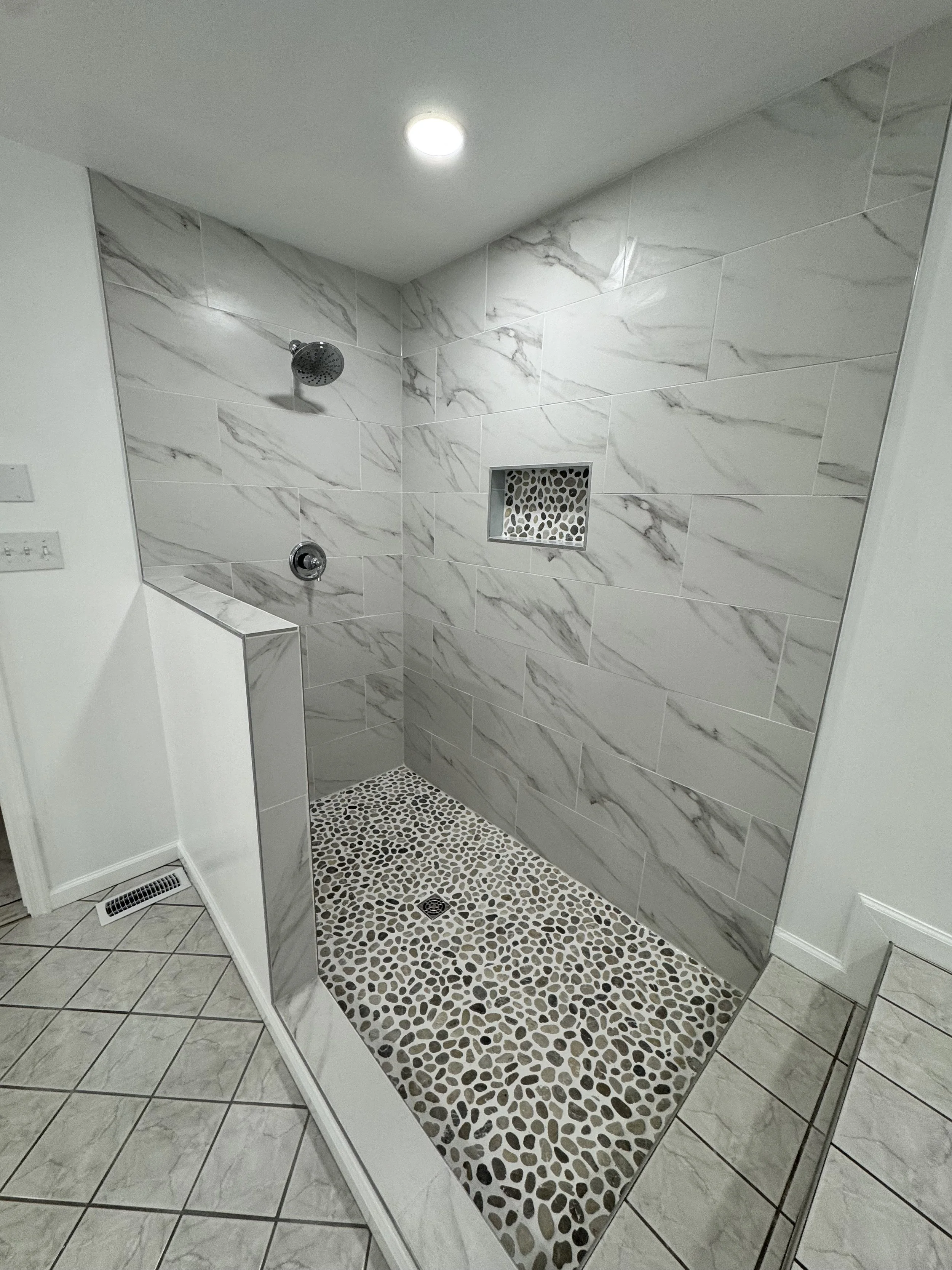 A modern shower with white marble wall tiles, a pebble mosaic floor, a built-in niche, a showerhead, and a single ceiling light.