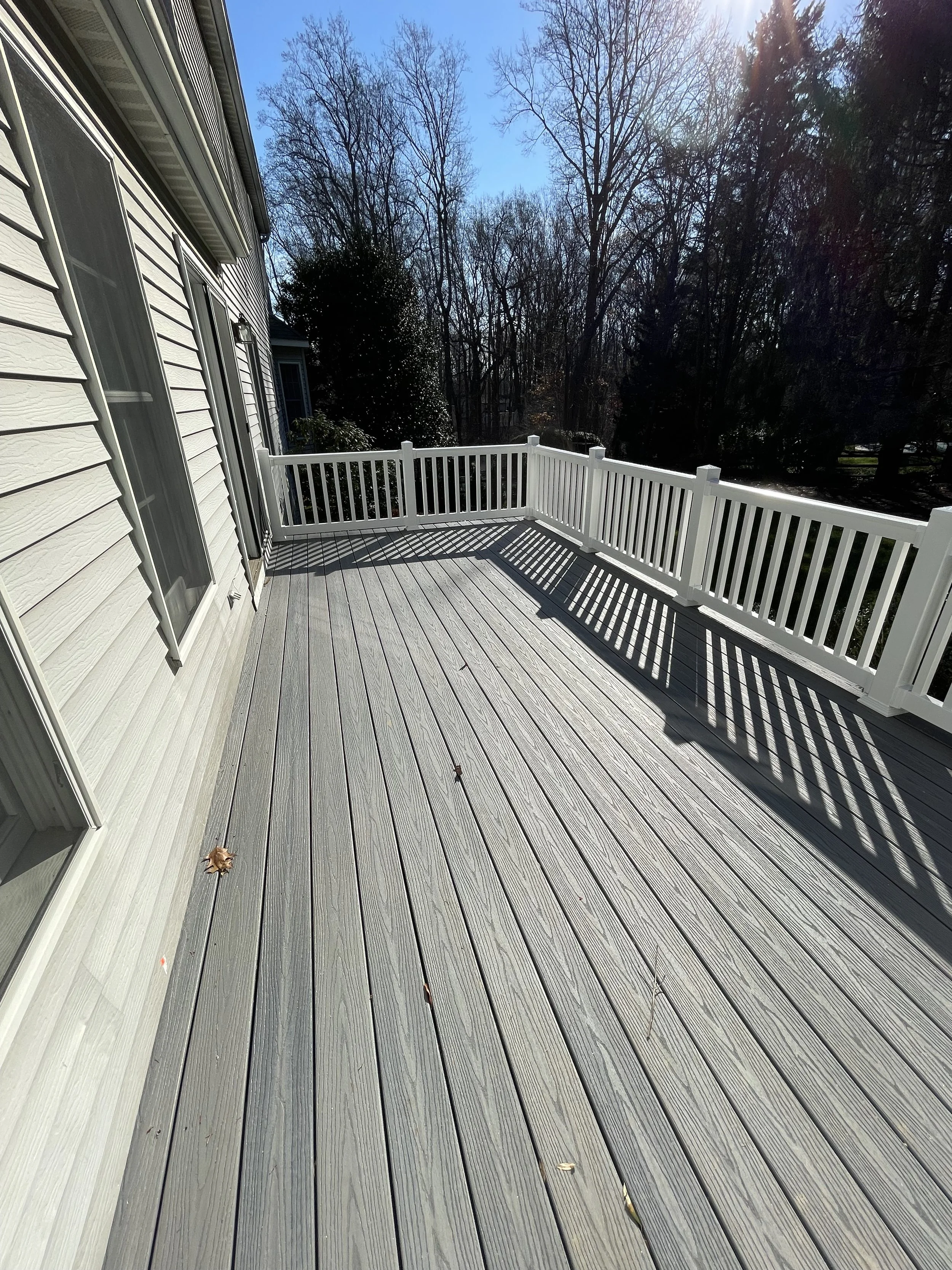 Empty outdoor deck with white railing, gray wooden floor, and leaf shadows, adjacent to a house with white siding, under a clear blue sky.