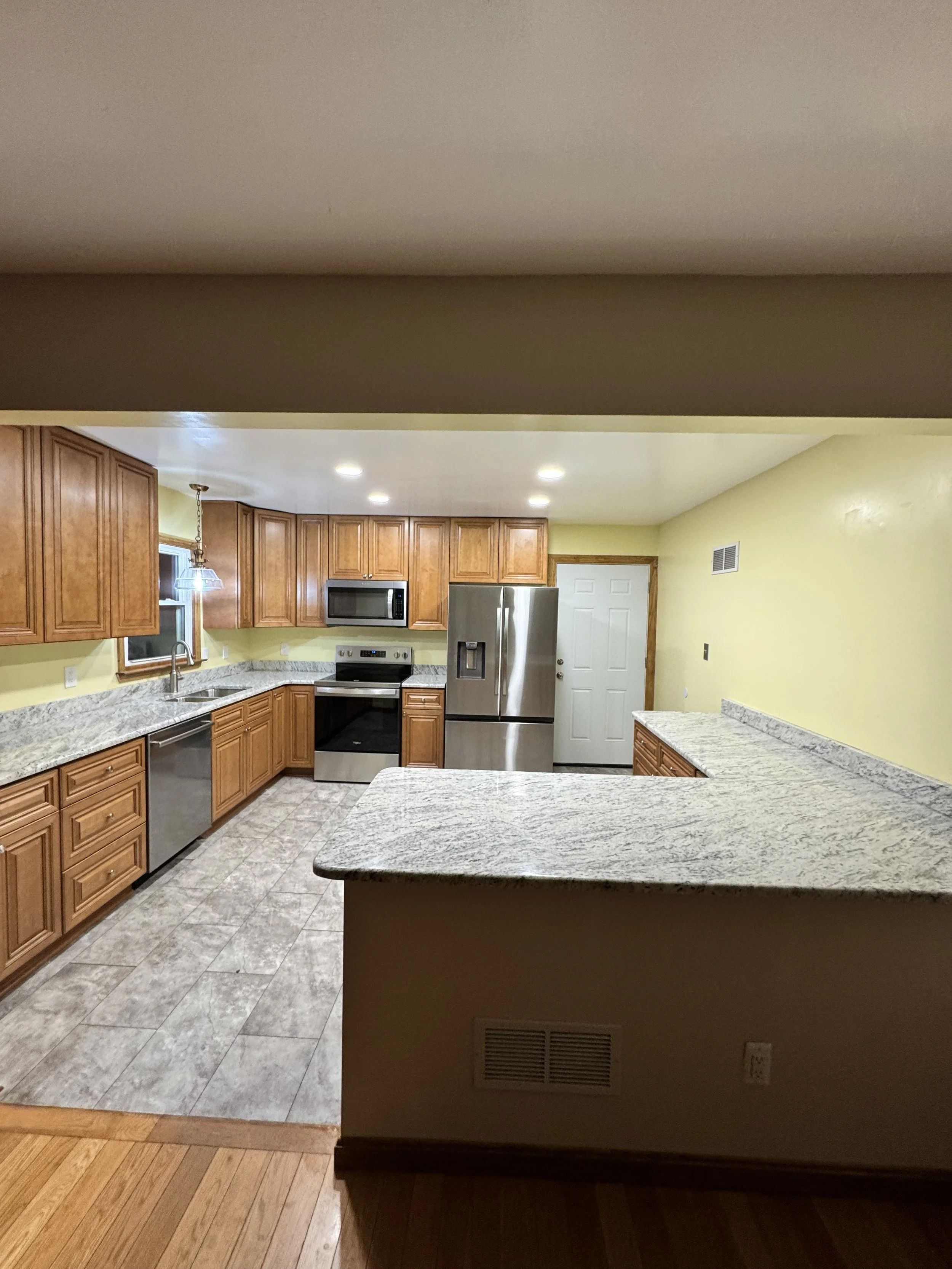 Kitchen with wooden cabinets, granite countertops, stainless steel appliances, including a refrigerator, microwave, and oven, yellow walls, tiled floor, and recessed lighting.