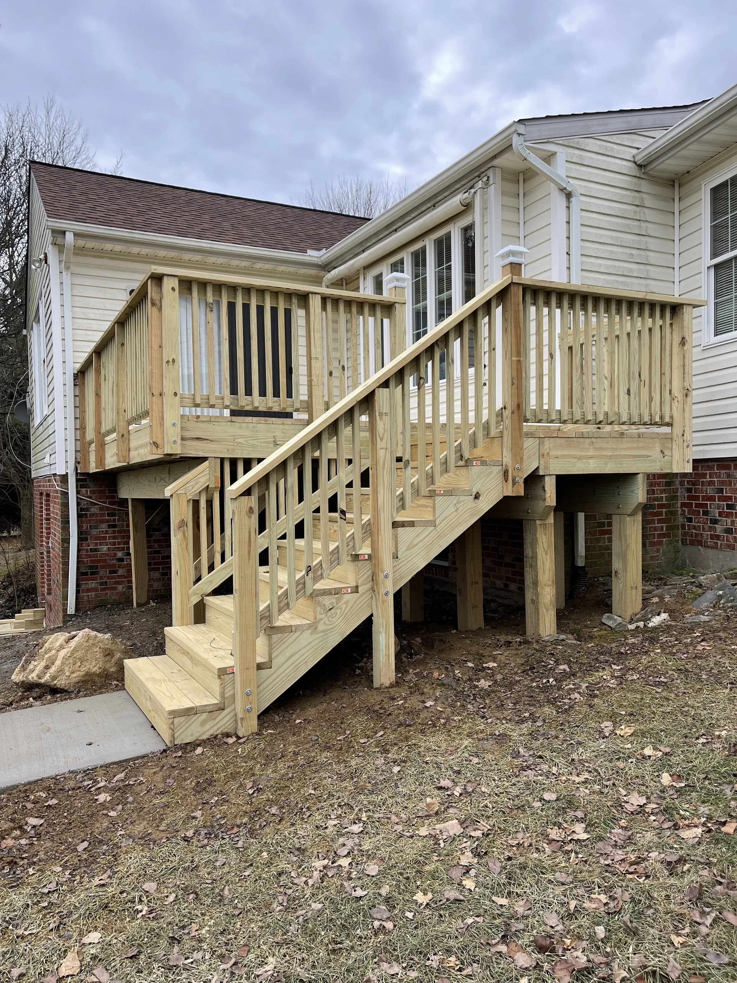 Unfinished wooden deck with stairs attached to the back of a white house, with a cloudy sky in the background.