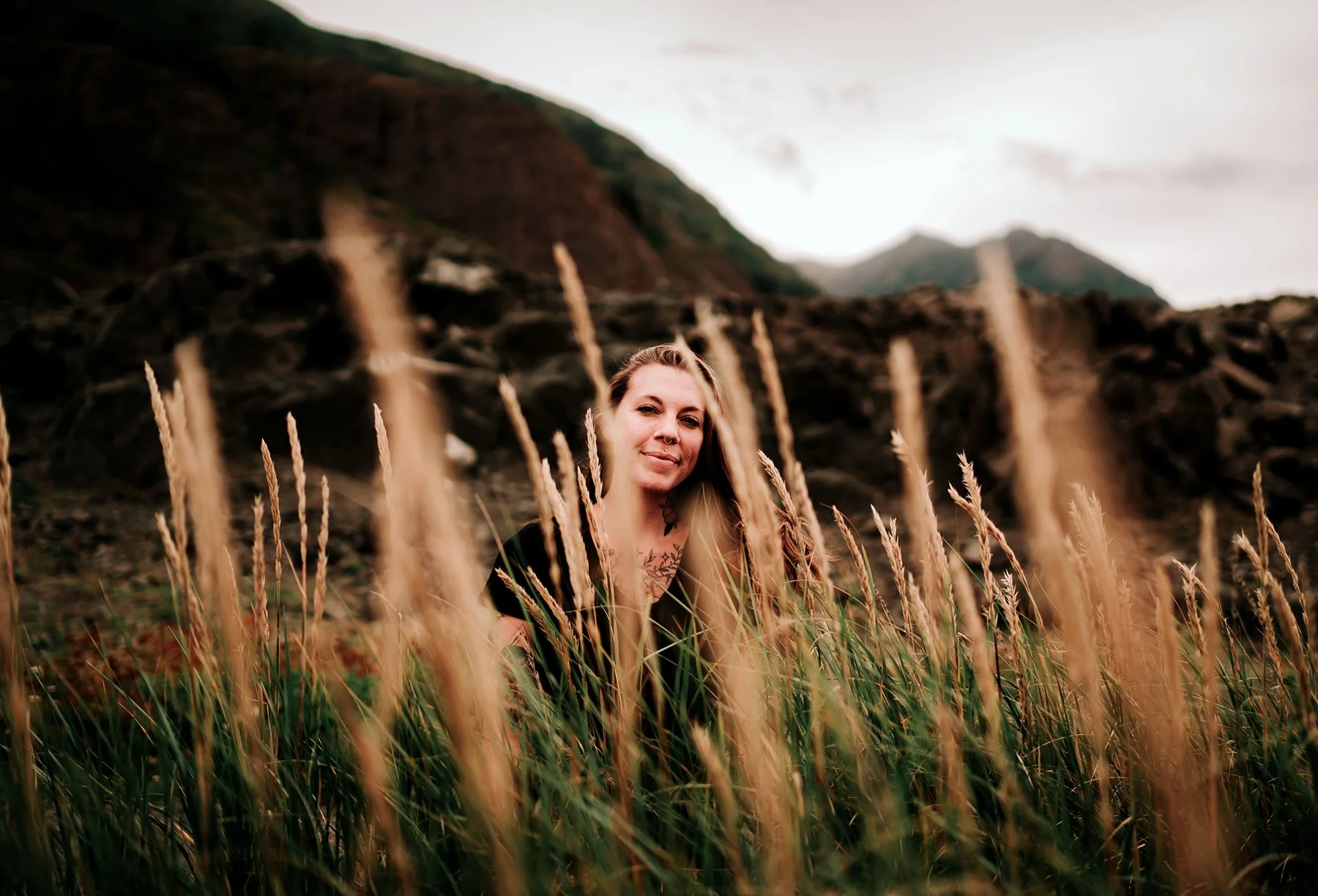 A woman with long blonde hair and tattoos on her chest, smiling behind tall grass in a mountainous landscape with overcast sky.